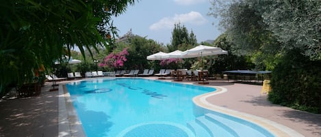 Una piscina con sillones de descanso y sombrillas, rodeada de vegetación y árboles.