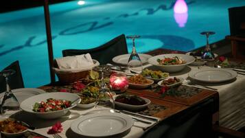 A dining table set for a meal with various dishes and glasses, overlooking a pool with a blue hue.