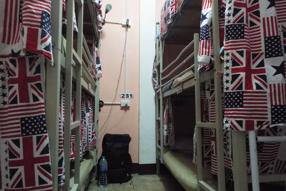 A narrow corridor with bunk beds on both sides, each bed covered with a blanket featuring the American and British flags. A black backpack and a water bottle are placed on the floor.