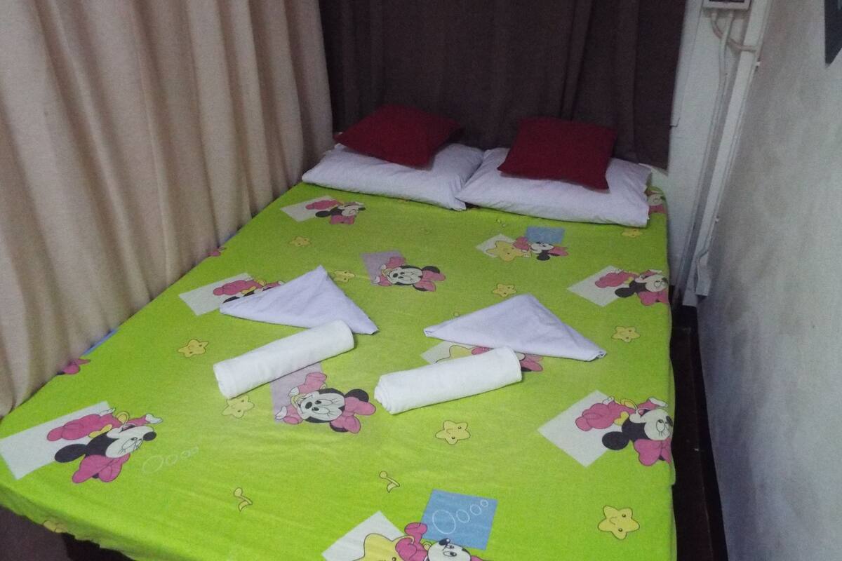 A single bed with a green bedspread featuring cartoon characters, white pillows, and neatly folded white towels.