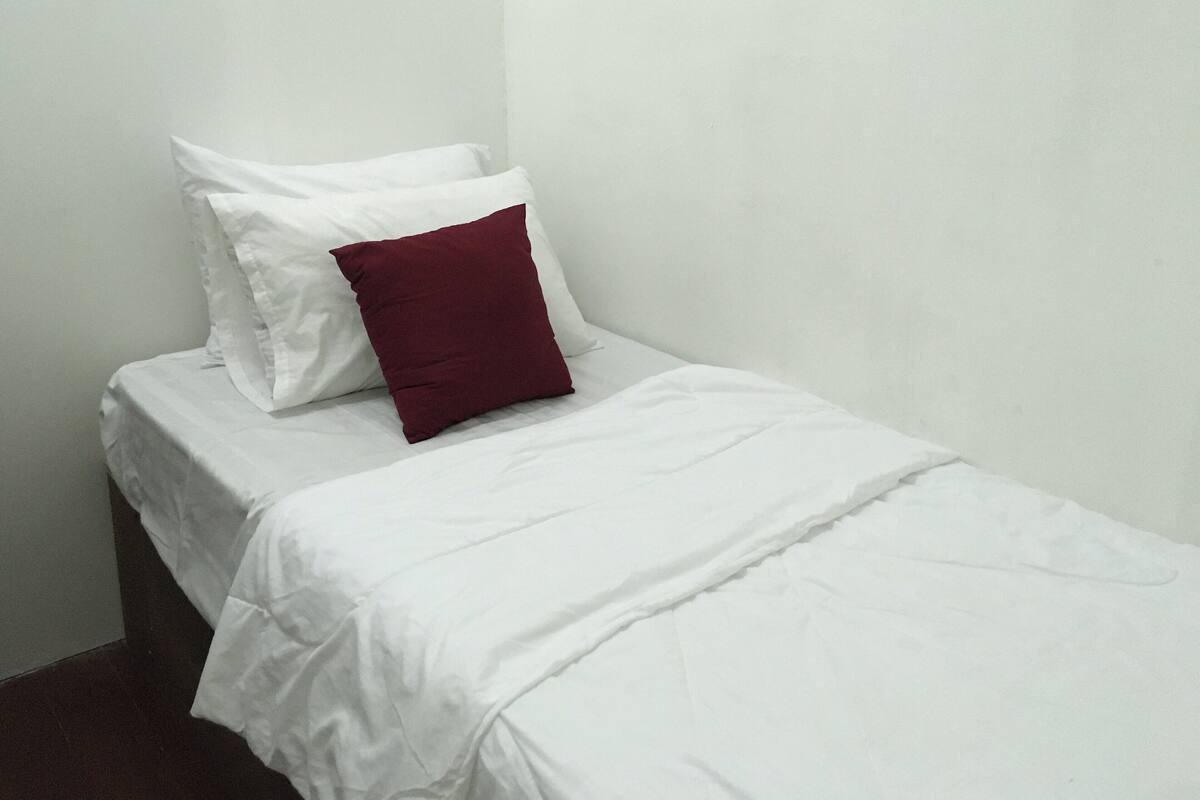 A small, single-bed room with white bedding and a red accent pillow.