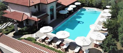 A swimming pool with sun loungers and umbrellas, surrounded by greenery and a stone building with a tiled roof.