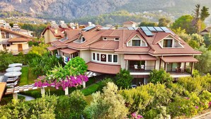 Une grande maison à plusieurs étages, dotée de panneaux solaires sur le toit, entourée d’une végétation luxuriante et avec une chaîne de montagnes en toile de fond.