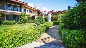 Un chemin menant à un bâtiment doté d’un balcon, entouré de haies et d’arbres bien entretenus.