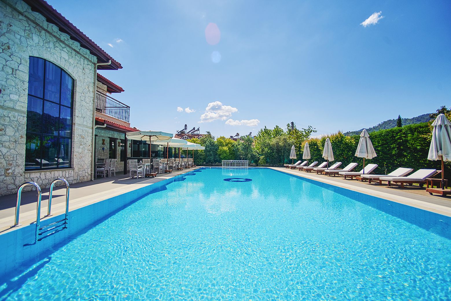 Una piscina azul cristalina con sillas de descanso y sombrillas, rodeada de edificios de piedra y follaje.