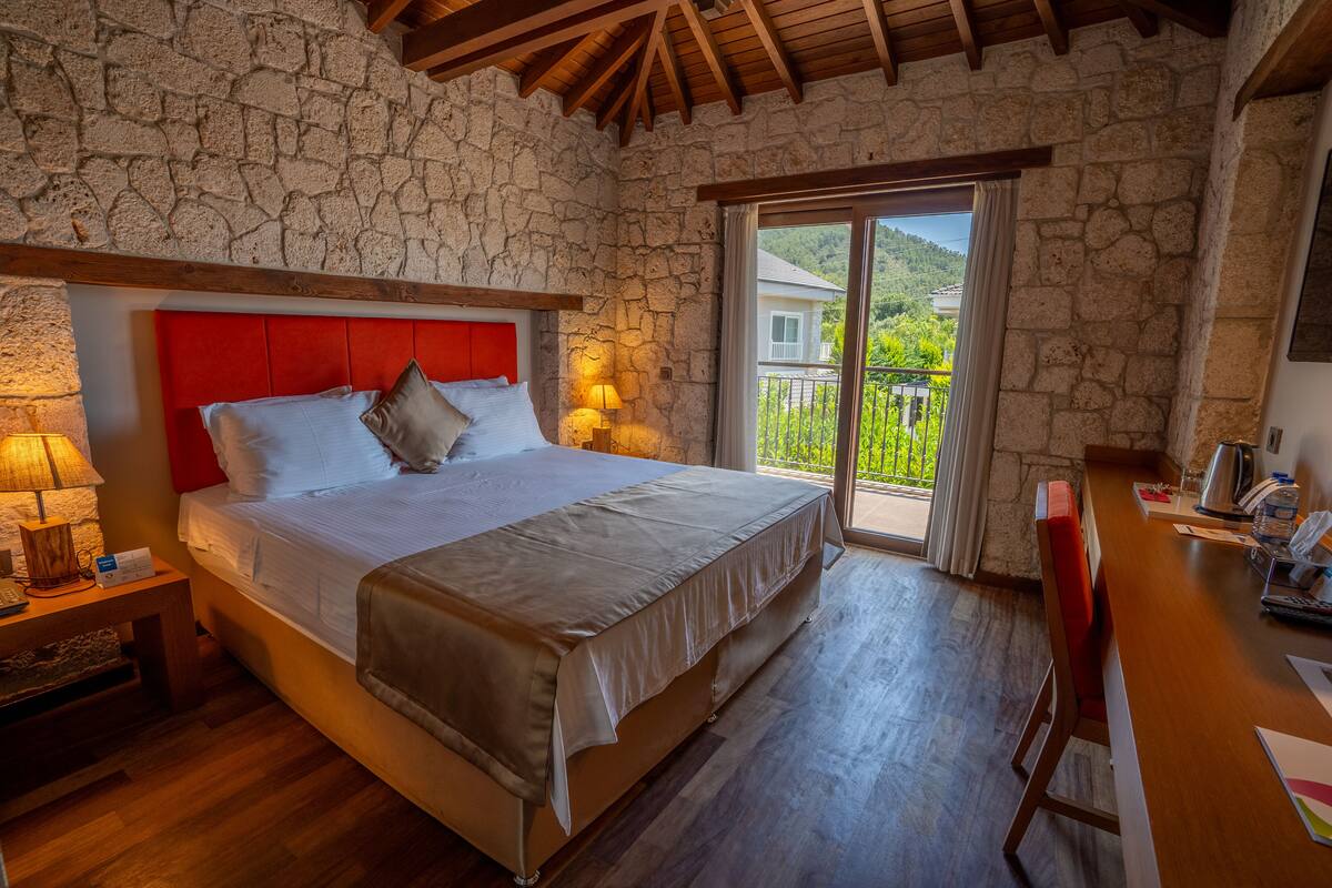 Une chambre avec un mur en pierre, un plafond en bois, un lit avec une tête de lit rouge, une table de chevet avec une lampe, et une vue sur la verdure à l’extérieur.