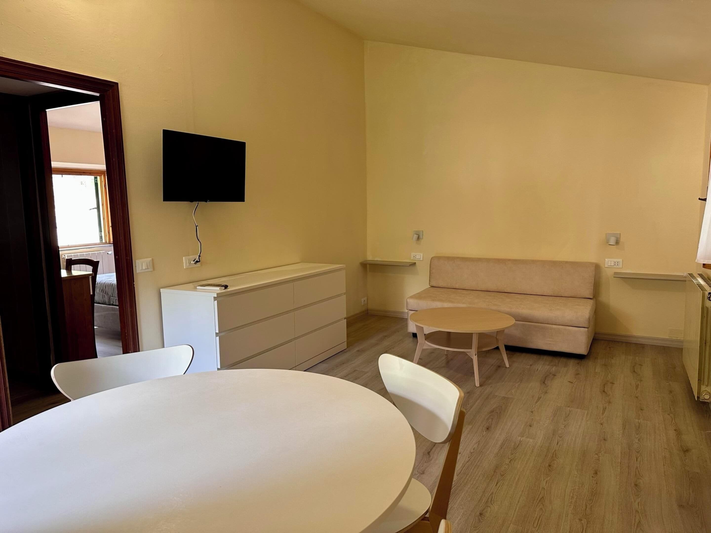 A living room with a white sofa, a small round table, and a white chair. There is a TV mounted on the wall and a wooden cabinet.