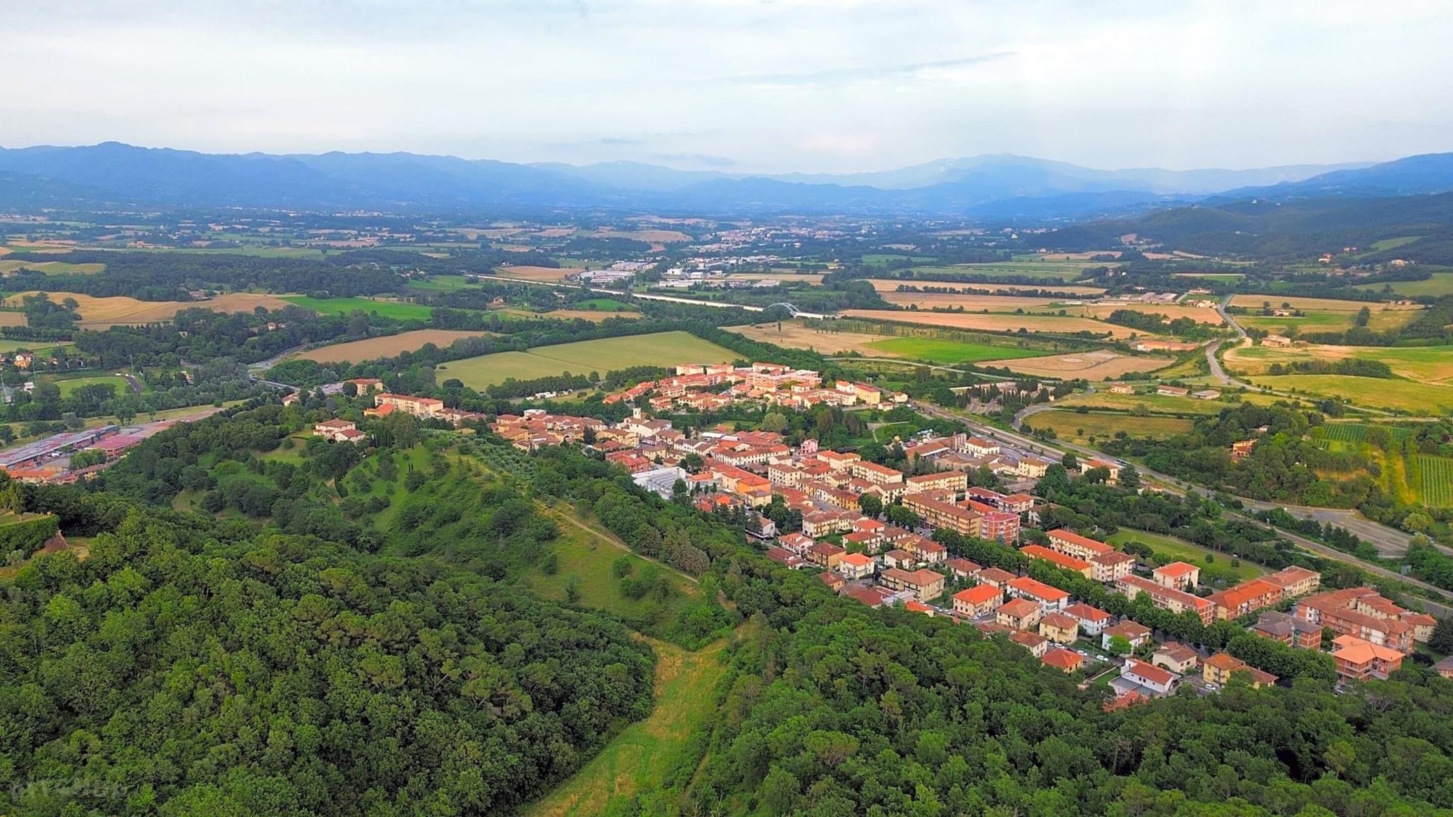 Vista aérea de uma cidade com um conjunto denso de edifícios com telhados vermelhos, rodeada por campos verdes e montanhas ao fundo.