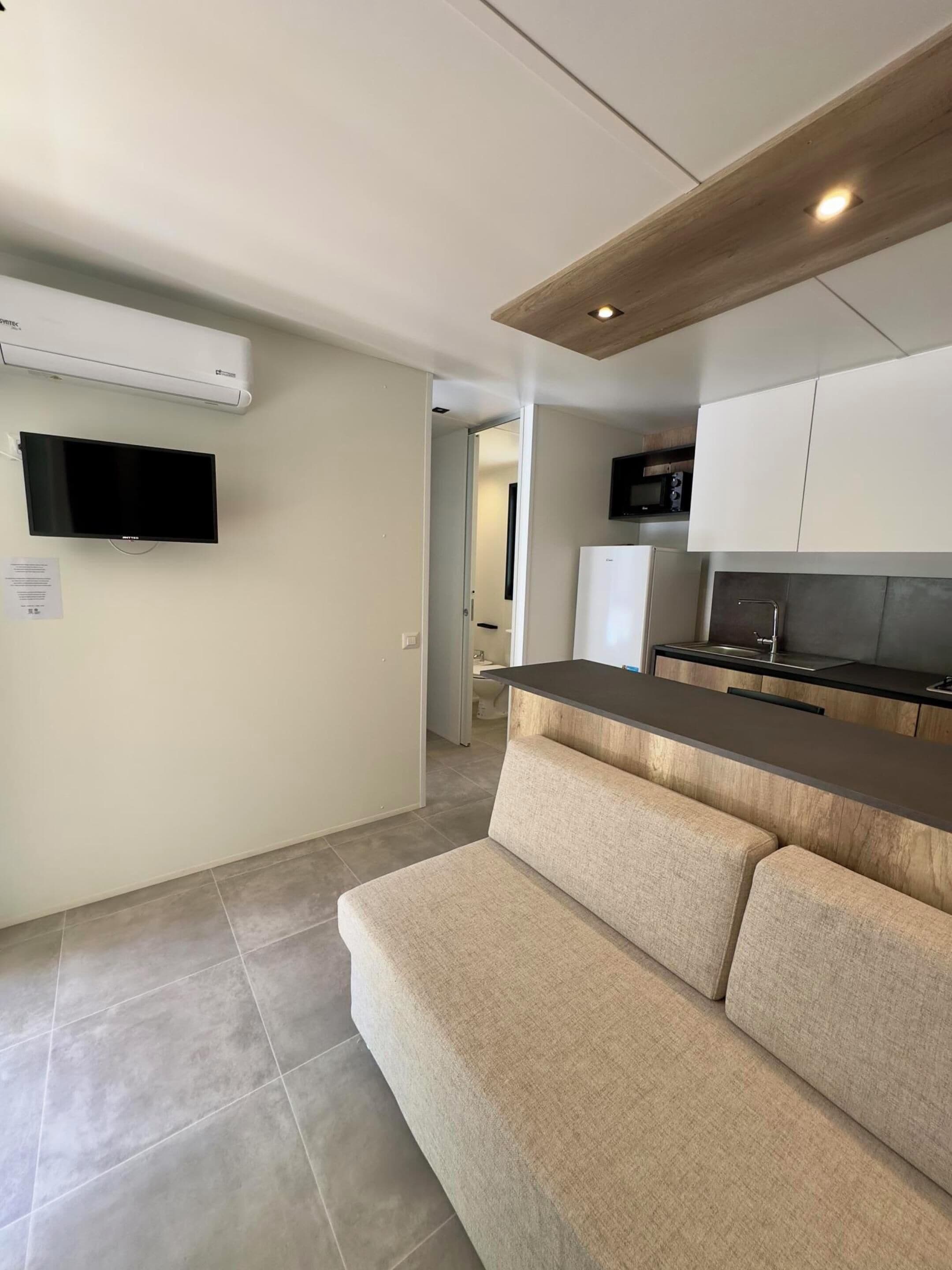 Una cocina moderna con horno empotrado, microondas y lavamanos. Hay un sofá color beige y un televisor de pantalla plana montado en la pared.