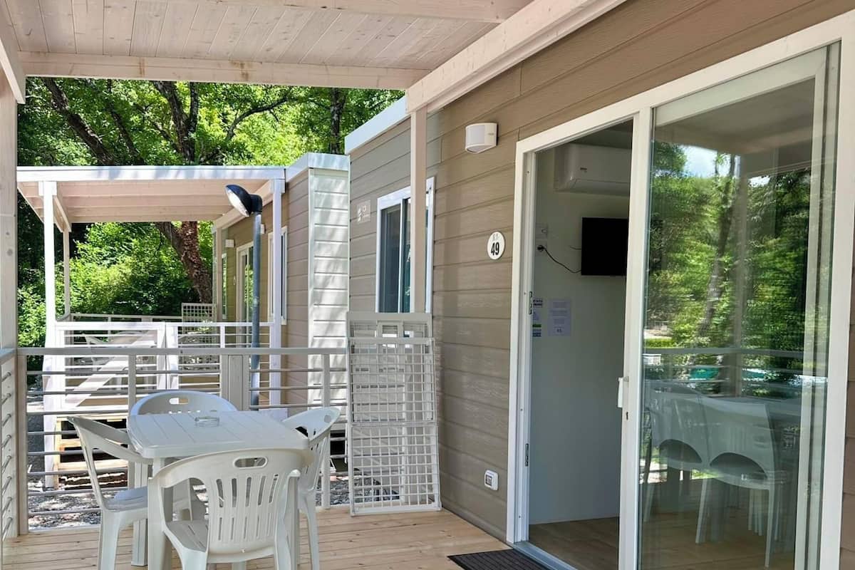 Zona exterior coberta amb terrassa de fusta, taula i cadires, i una porta corredissa de vidre que dona a una habitació.