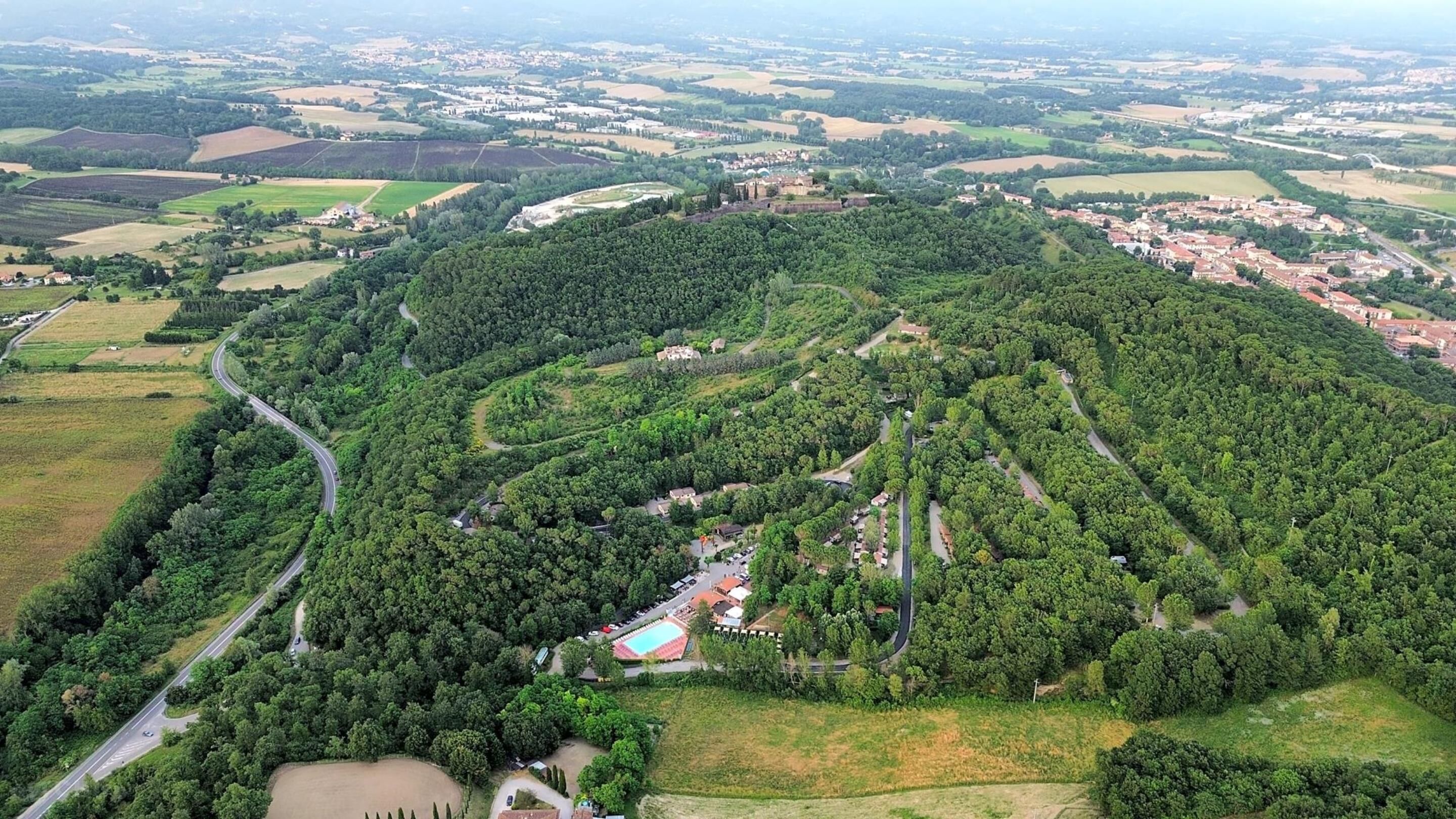An aerial view of a hilly area with a winding road, dense forest, and scattered buildings.