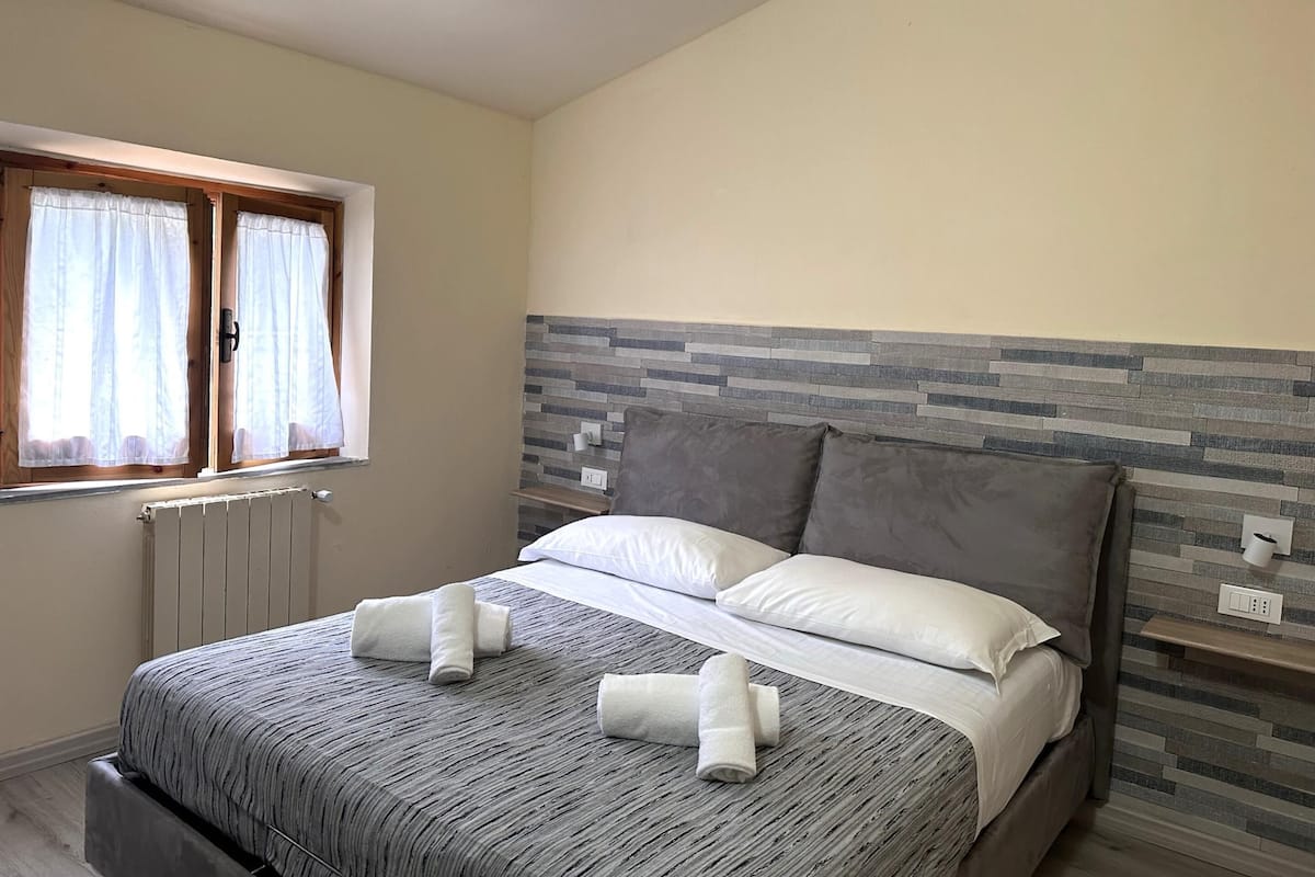 Un llit ben fet amb roba de llit gris i almoines blanques, una finestra amb cortines blanques i un panell de capçal gris i blanc amb dibuixos.