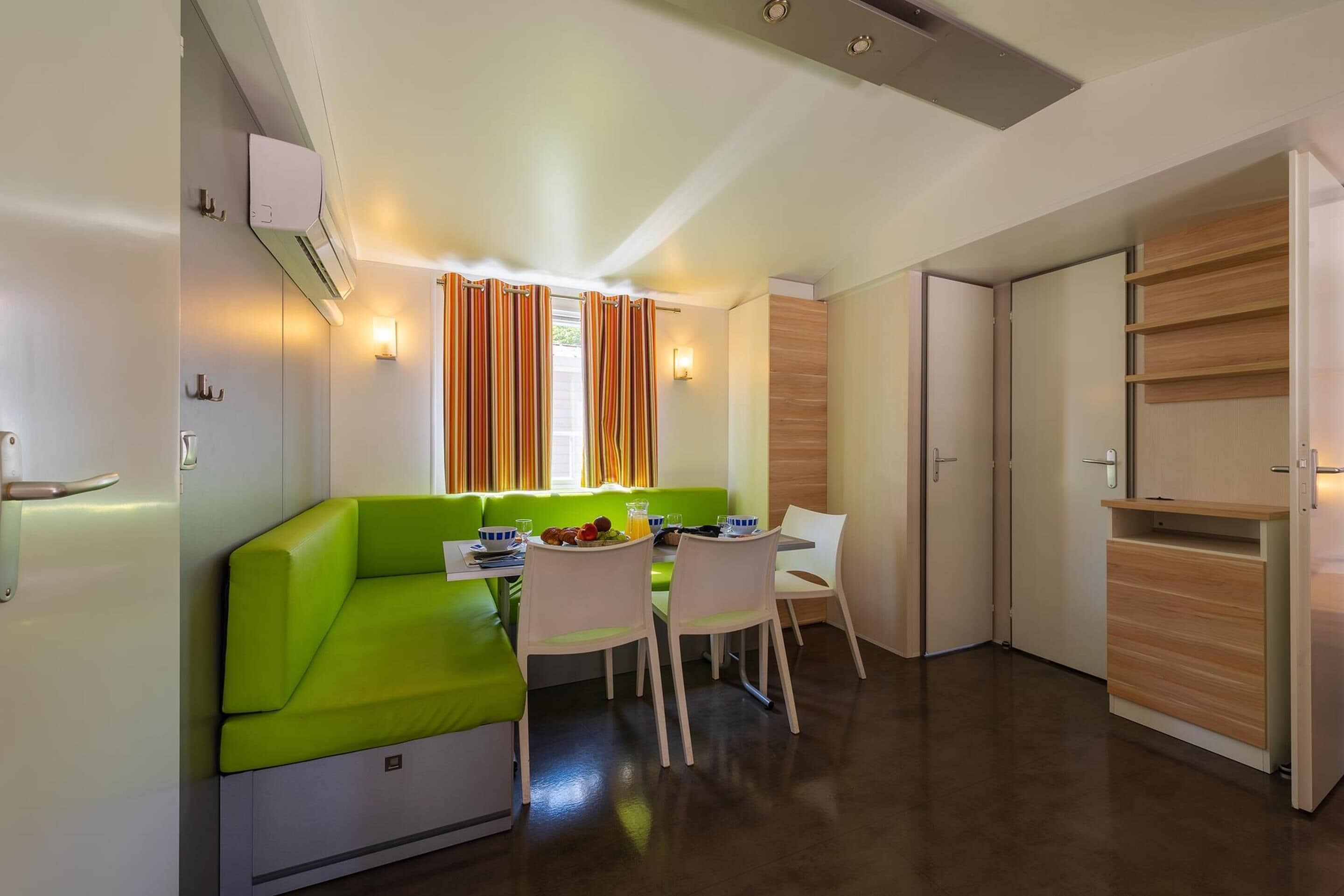 A compact living space with a green sofa, a dining table, and chairs. There is a kitchenette with a microwave and a small refrigerator. The room has a window with curtains and a wall-mounted light fixture.