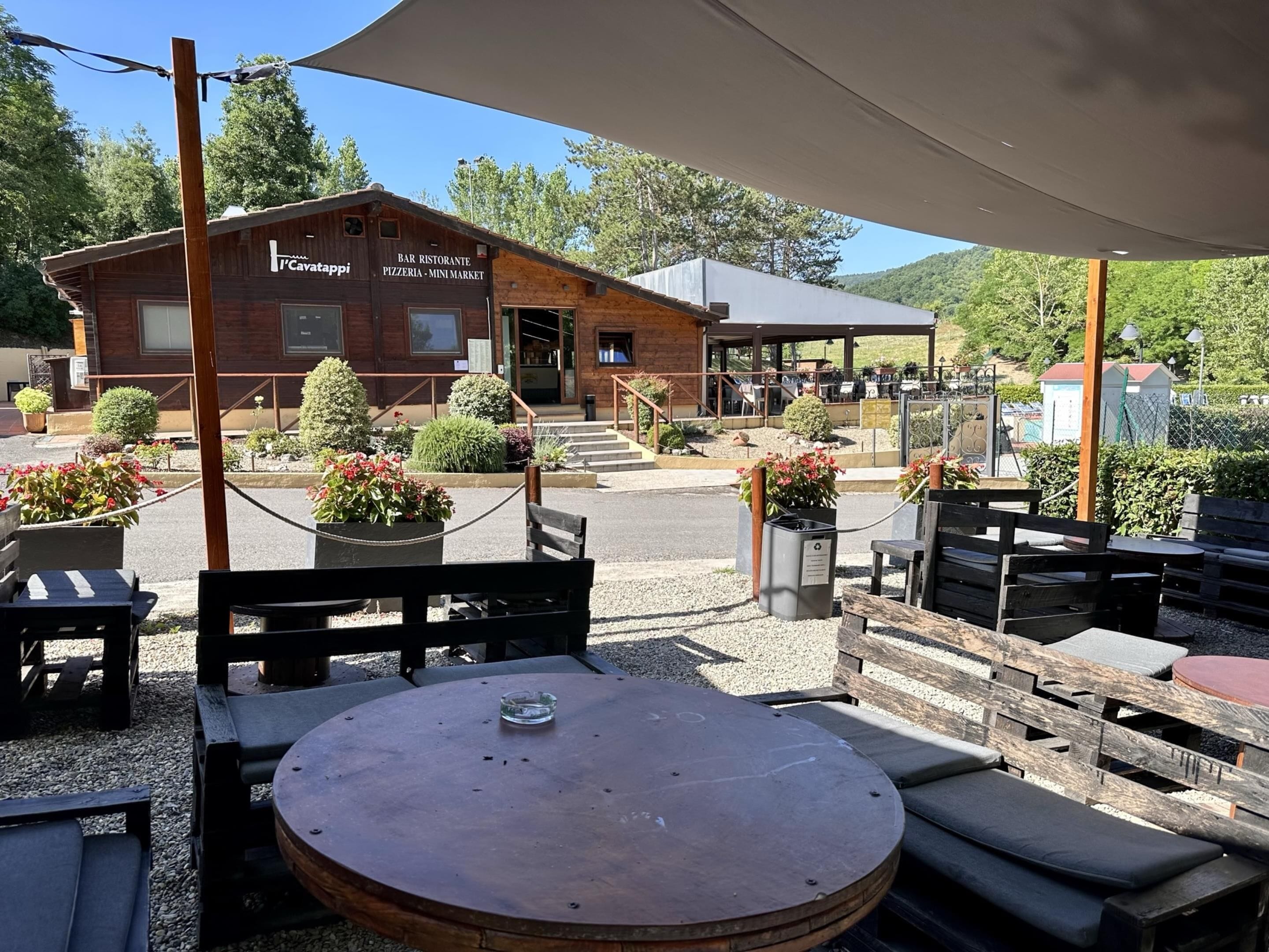 An outdoor seating area with wooden tables and benches, surrounded by potted plants and a building labeled "Bar Ristorante Pizzeria - Mini Market".