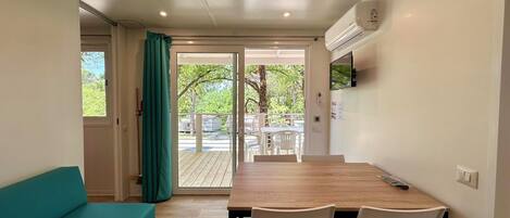 Una habitación compacta con una mesa de madera y sillas blancas, un sofá color turquesa y un ventanal corredero que da a un área exterior con terraza y sillas.