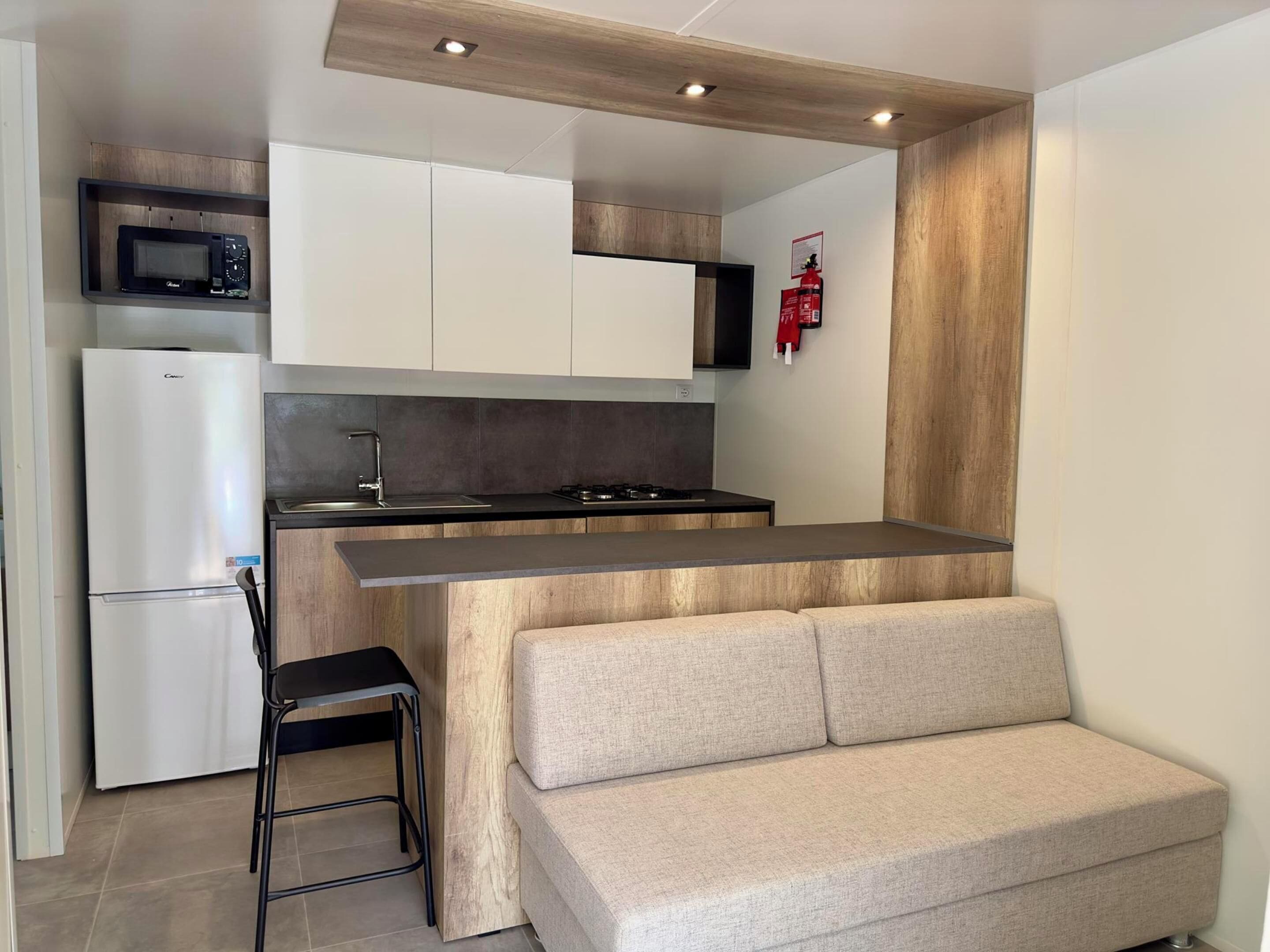 A compact kitchen with a microwave, refrigerator, and stove. There is a wooden accent wall and a small seating area with a sofa.