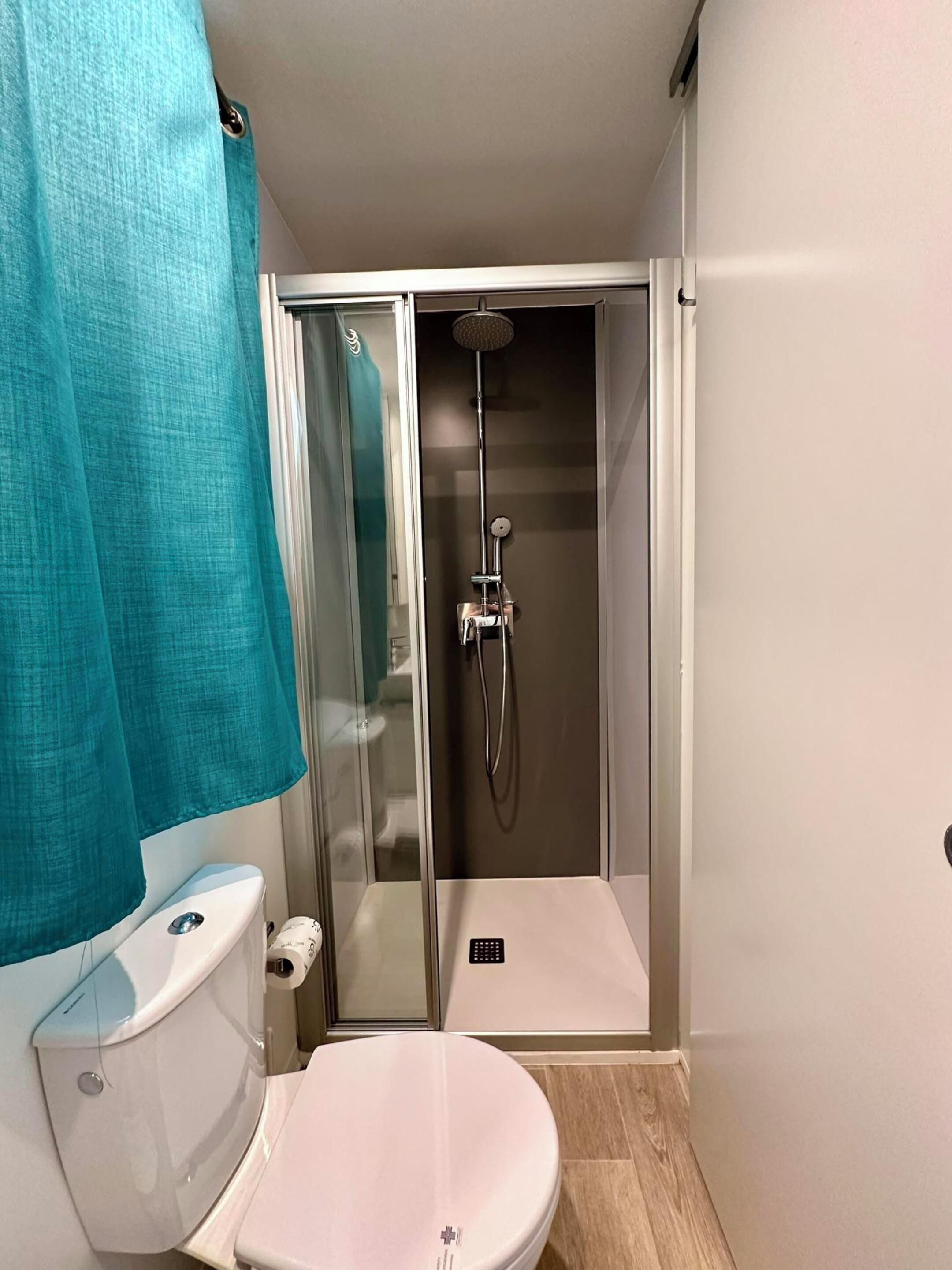 Un baño pequeño con inodoro, cabina de ducha con mampara de vidrio y cortina de ducha color verde azulado.