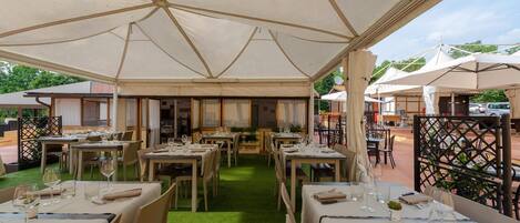 Un área de comedor al aire libre con mesas dispuestas para dos personas, cubierta por un toldo grande.