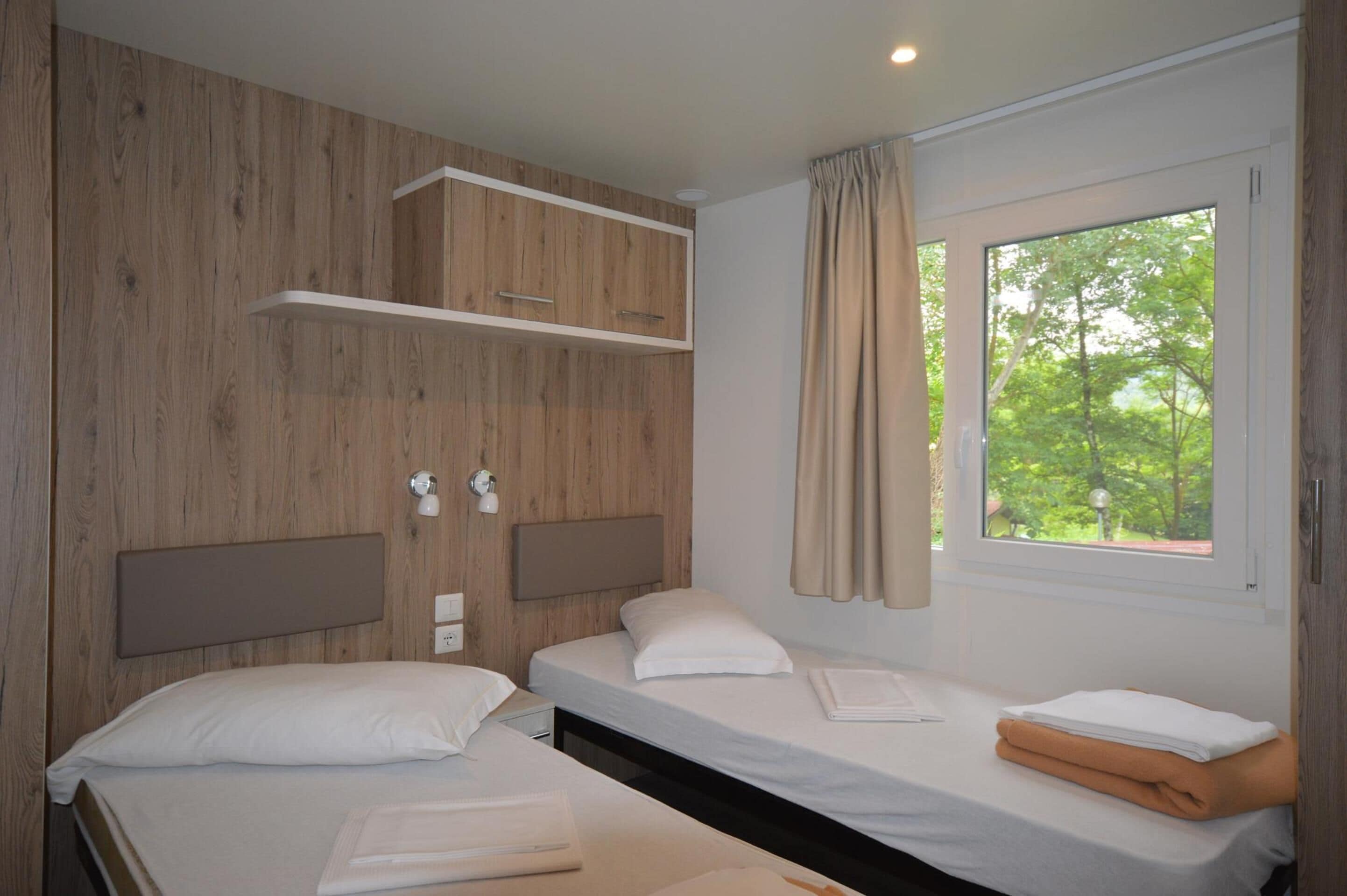 A bedroom with two single beds, wooden paneling on the walls, a window with curtains, and a small shelf above the beds.