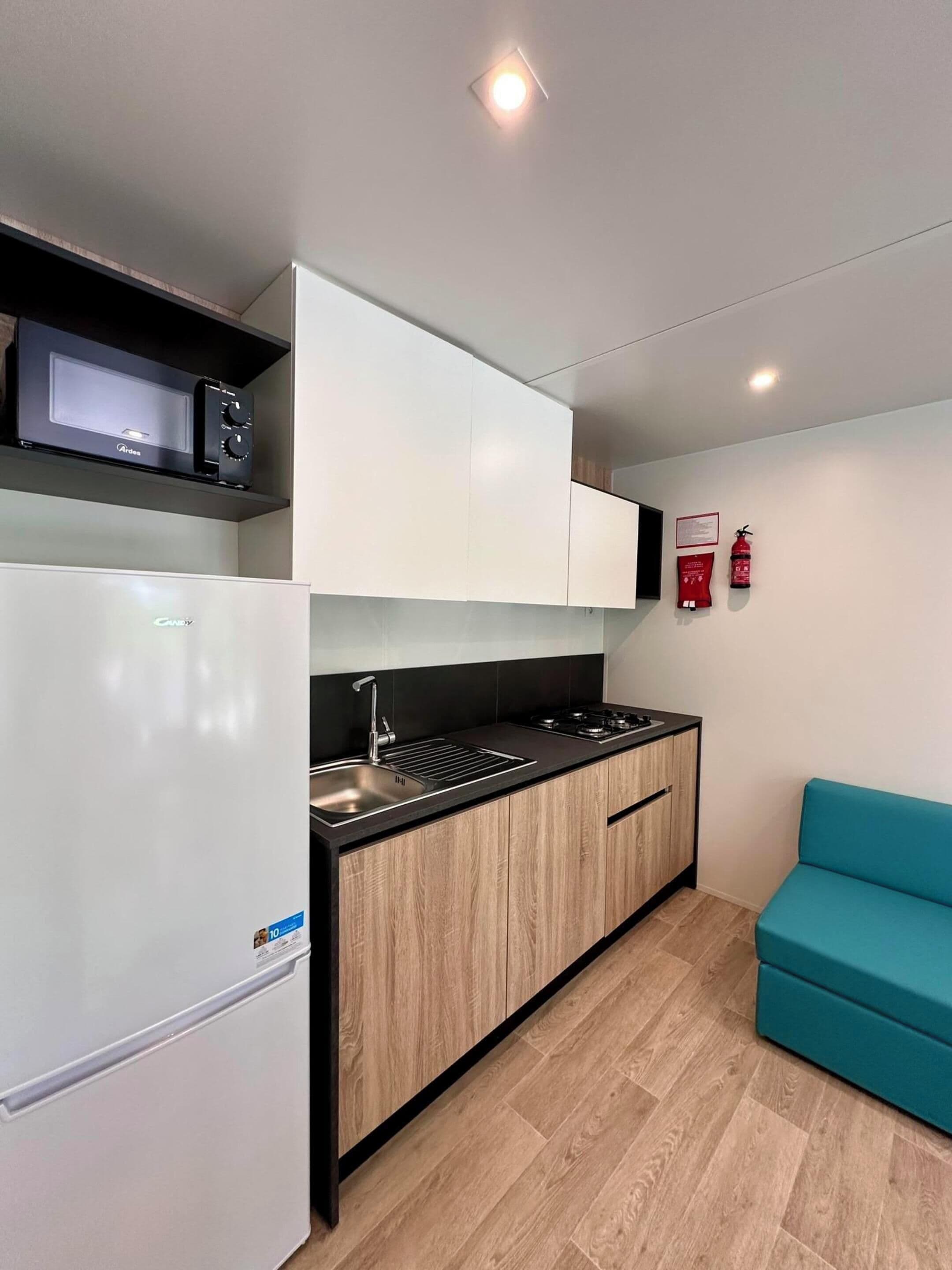 Cocina compacta con refrigerador, microondas, pileta y estufa. Hay un sofá color turquesa y un extintor de incendios en la pared.