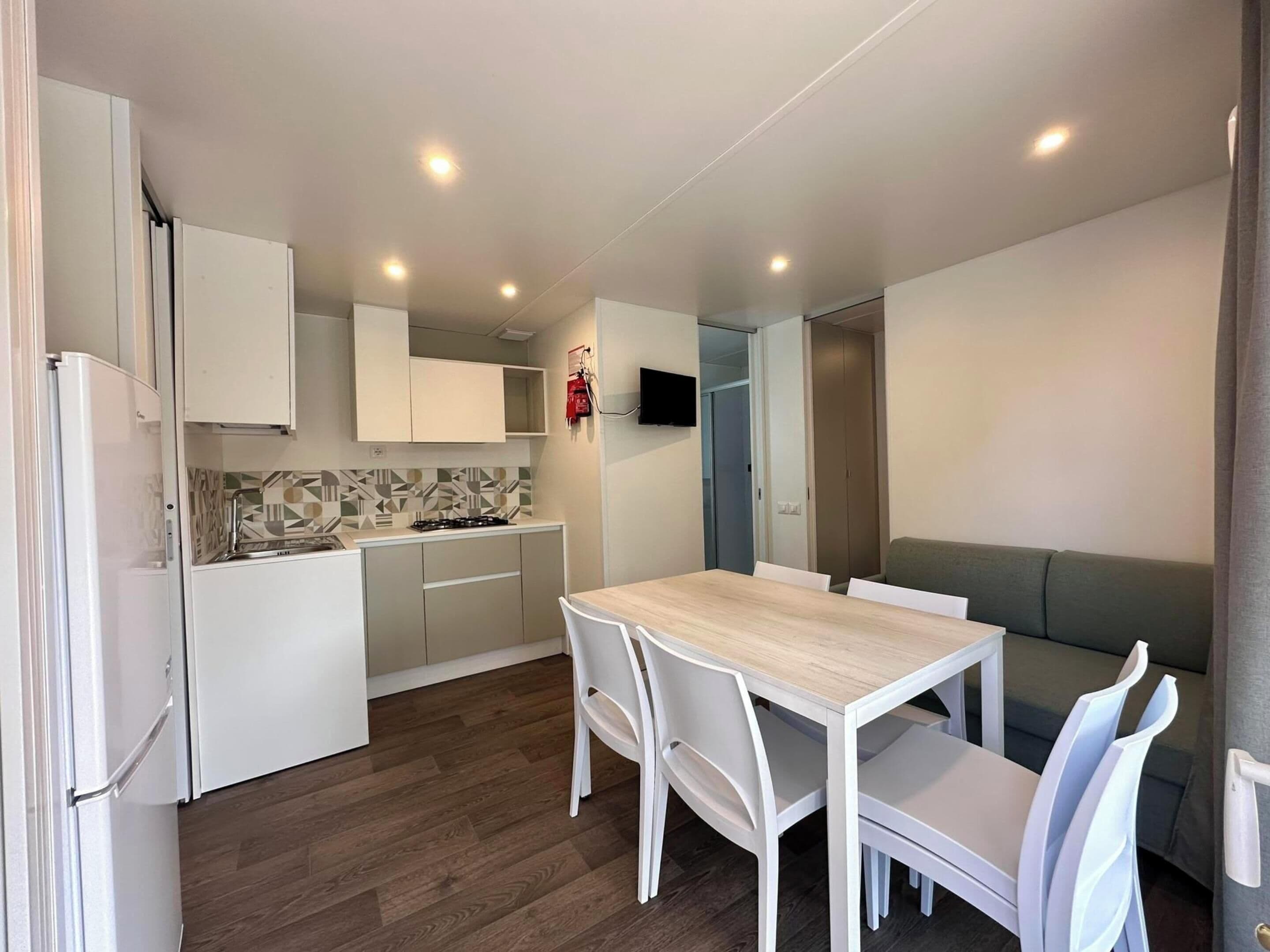 A compact kitchen with white cabinets, a tiled backsplash, and a built-in stove. There is a dining table with white chairs and a grey sofa.