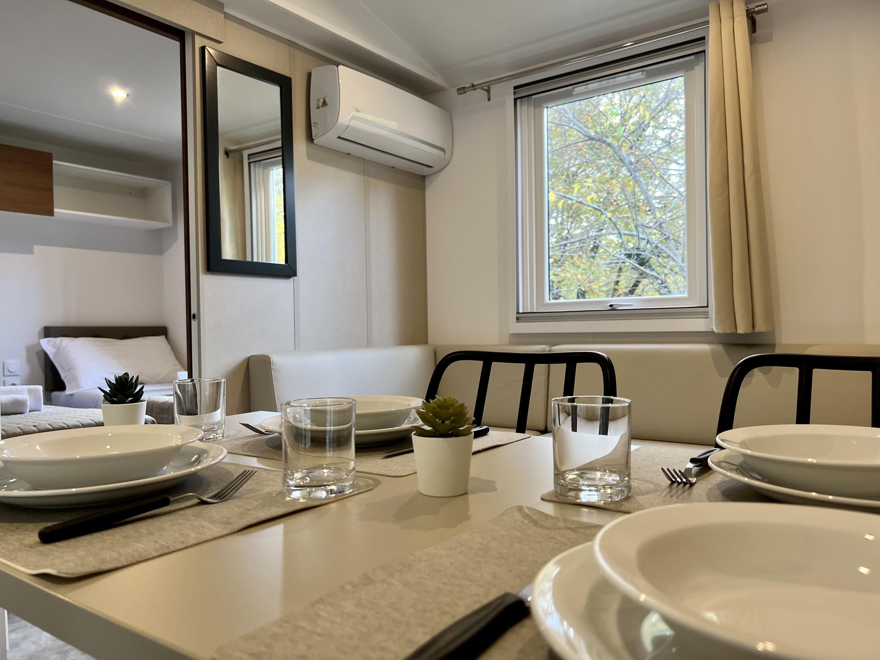 Un comedor ordenado con una mesa dispuesta para dos personas, una ventana con vista a árboles y un equipo de aire acondicionado en la pared.