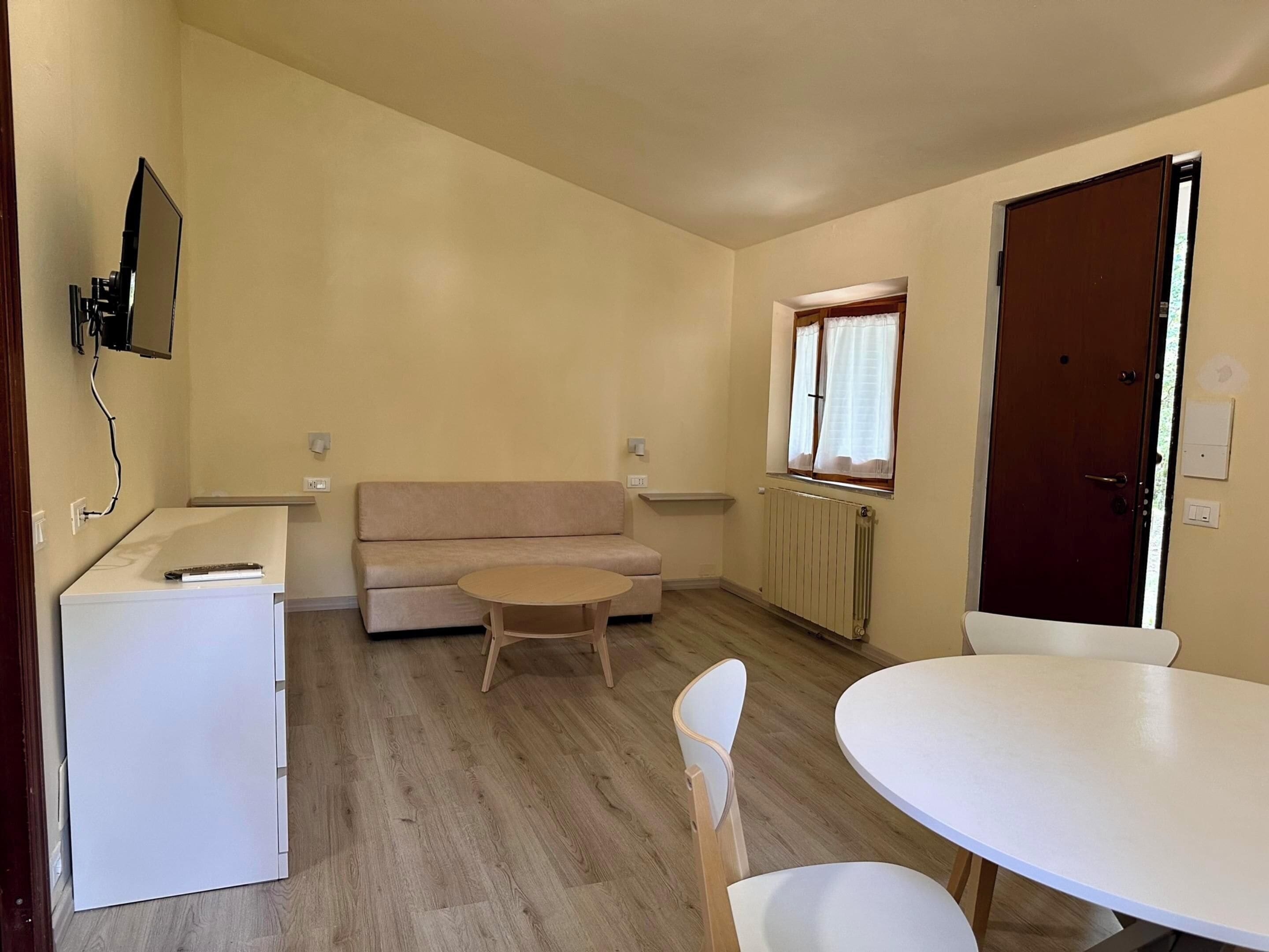 Un ambiente compatto arredato con un divano, un tavolino, una poltrona, una TV a parete e una porta.