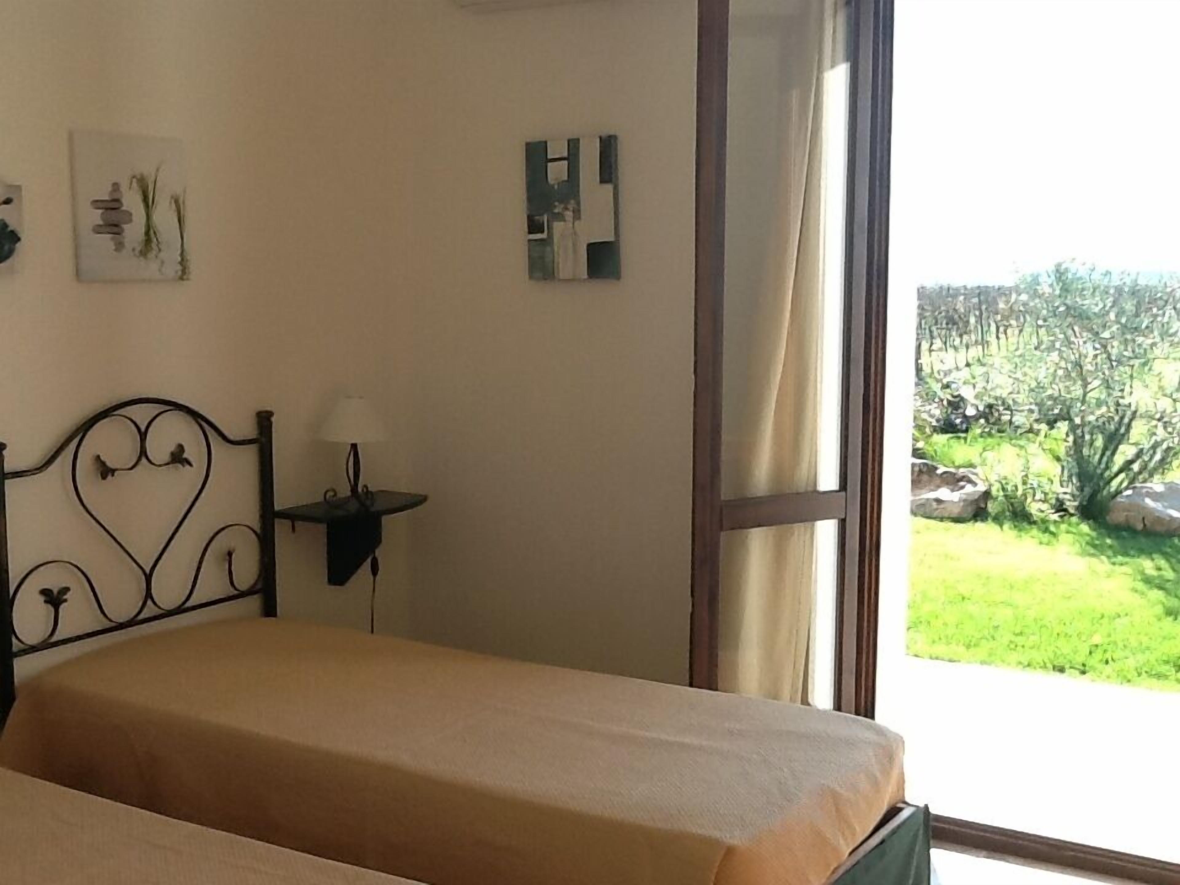 A bedroom with a bed, a bedside table with a lamp, a window with a view of a garden, and wall art.
