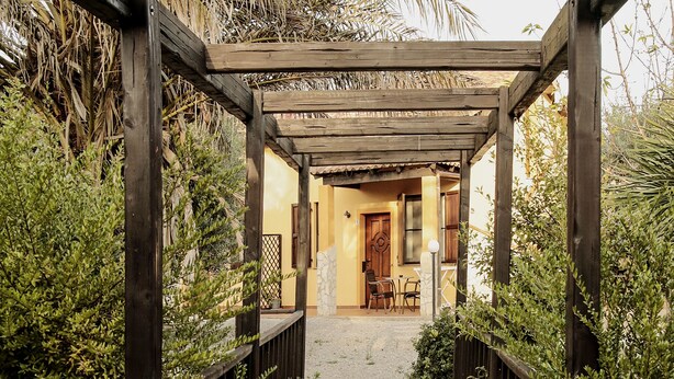 Un chemin de pierres mène à une pergola en bois surmontée d’un toit de chaume, le tout étant entouré d’une végétation luxuriante.