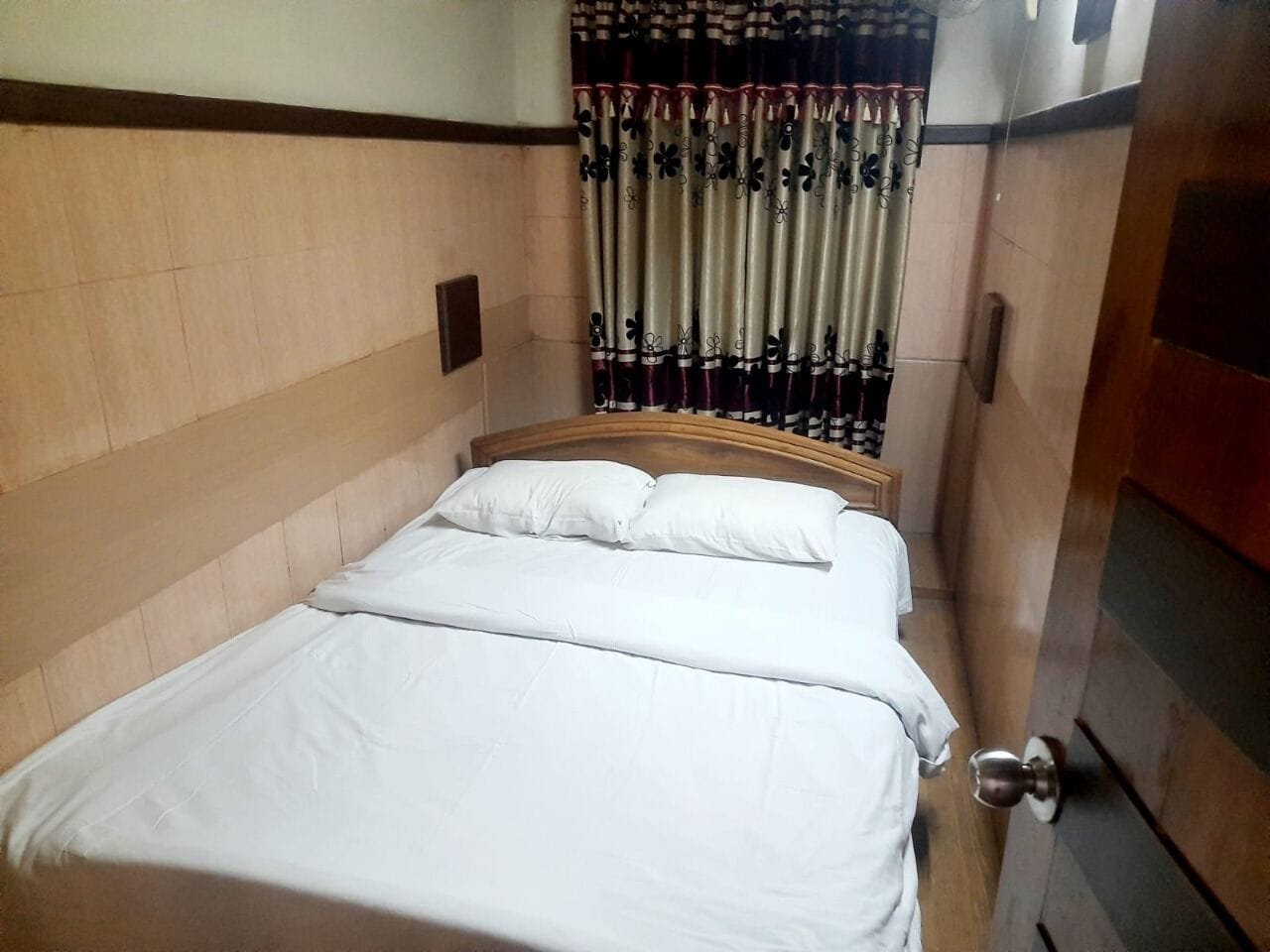 A neatly made bed with white linens in a room with wooden paneling on the walls and a patterned curtain.