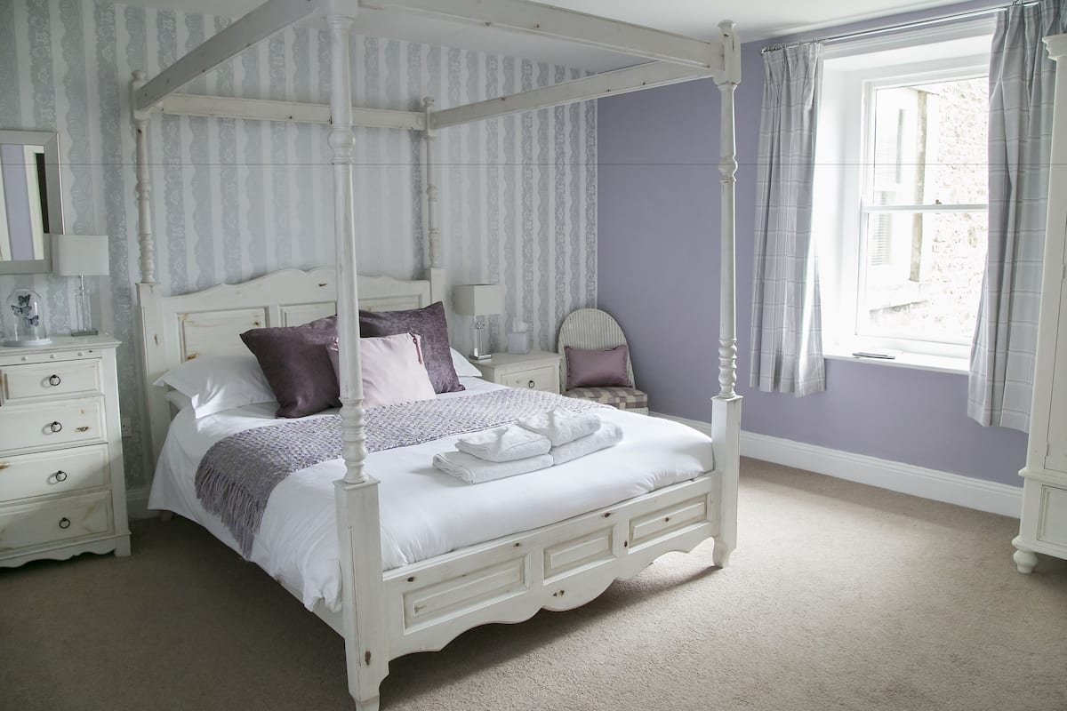 A four-poster bed with a white finish, a nightstand with a lamp, and a window with curtains.
