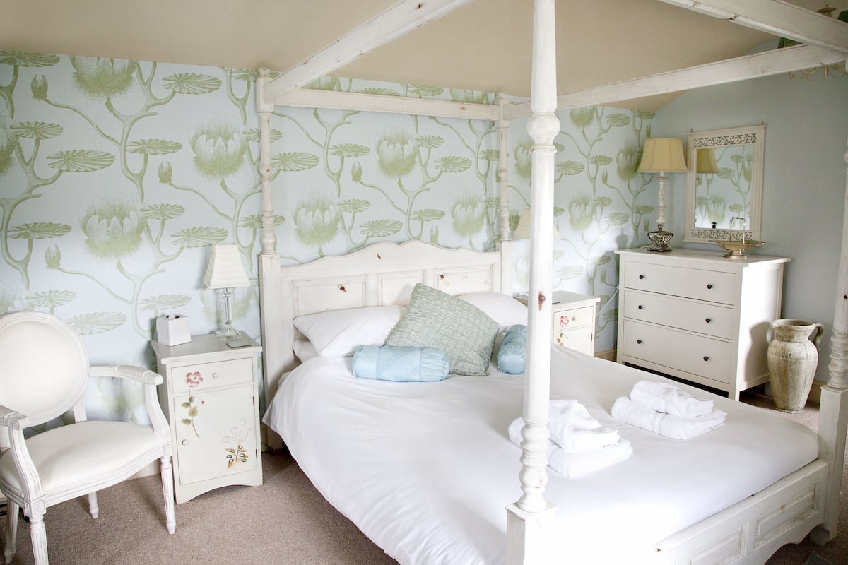 A four-poster bed with white bedding, a nightstand with a lamp, a dresser, and a chair in a room with floral wallpaper.