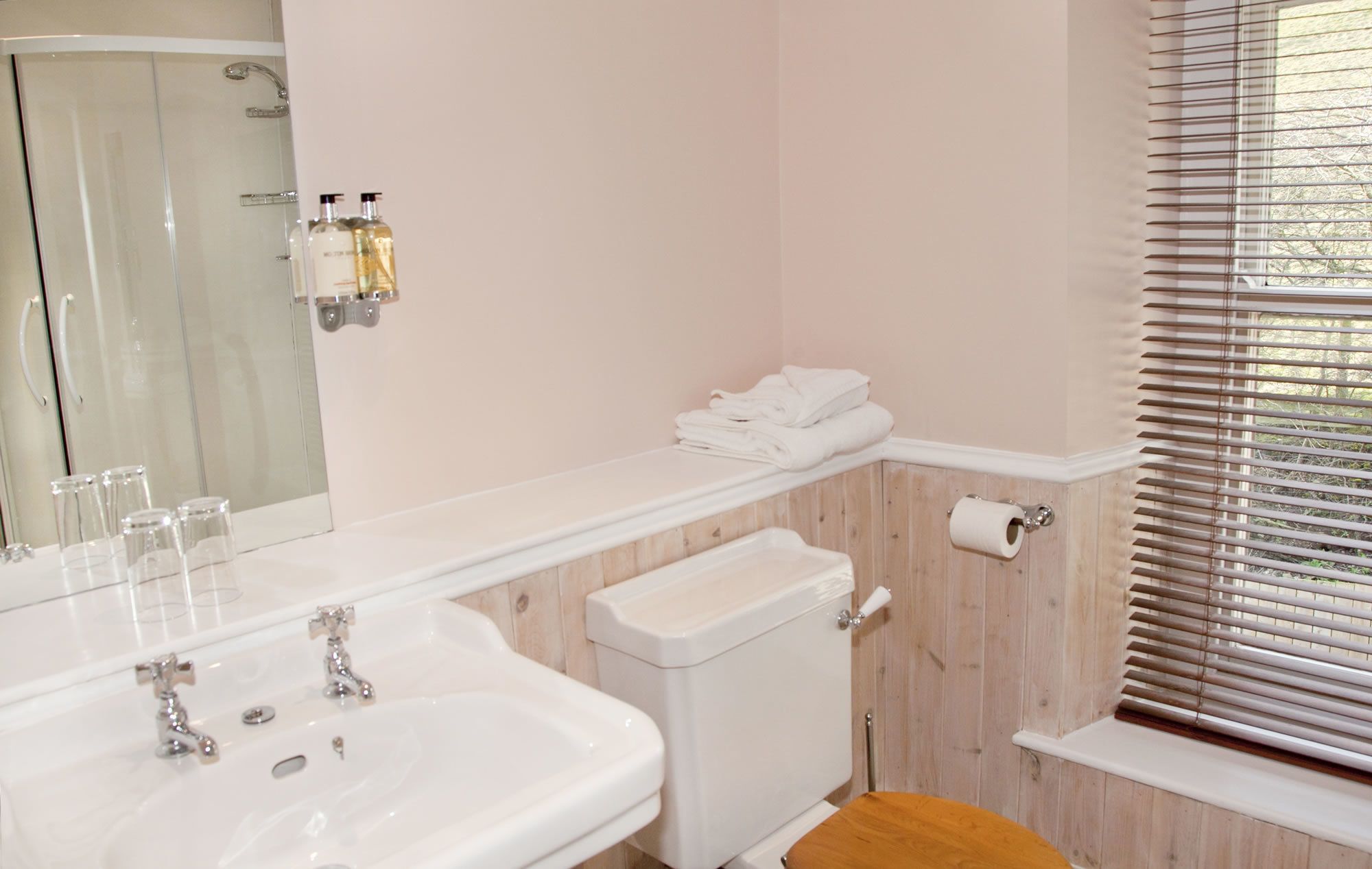 Une salle de bain avec un lavabo blanc, un miroir et des toilettes. Des serviettes sont pliées sur une étagère, et on aperçoit une douche au fond de la pièce.