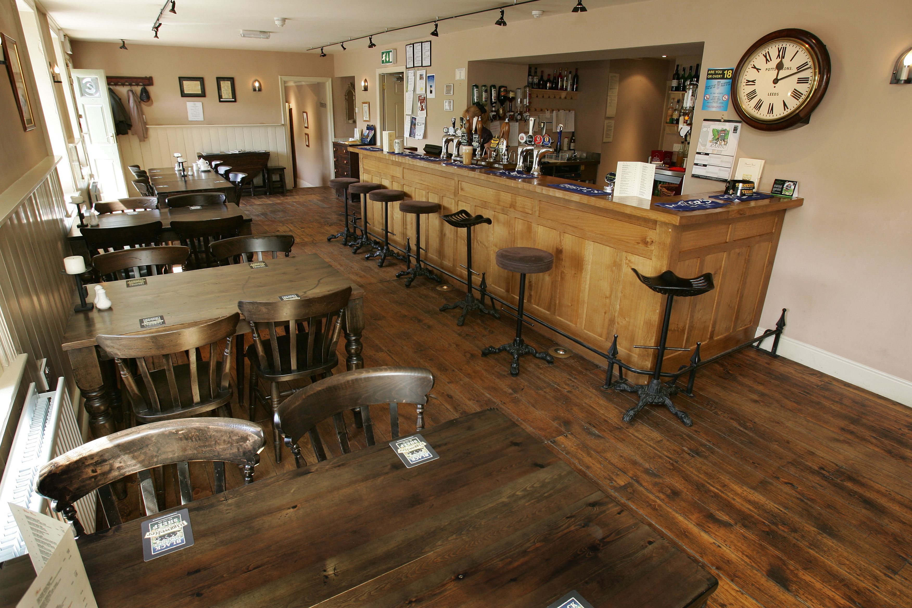 Un espace bar avec des tables et des tabourets en bois, un comptoir de bar orné d’une horloge, et un mur décoré de tableaux encadrés.