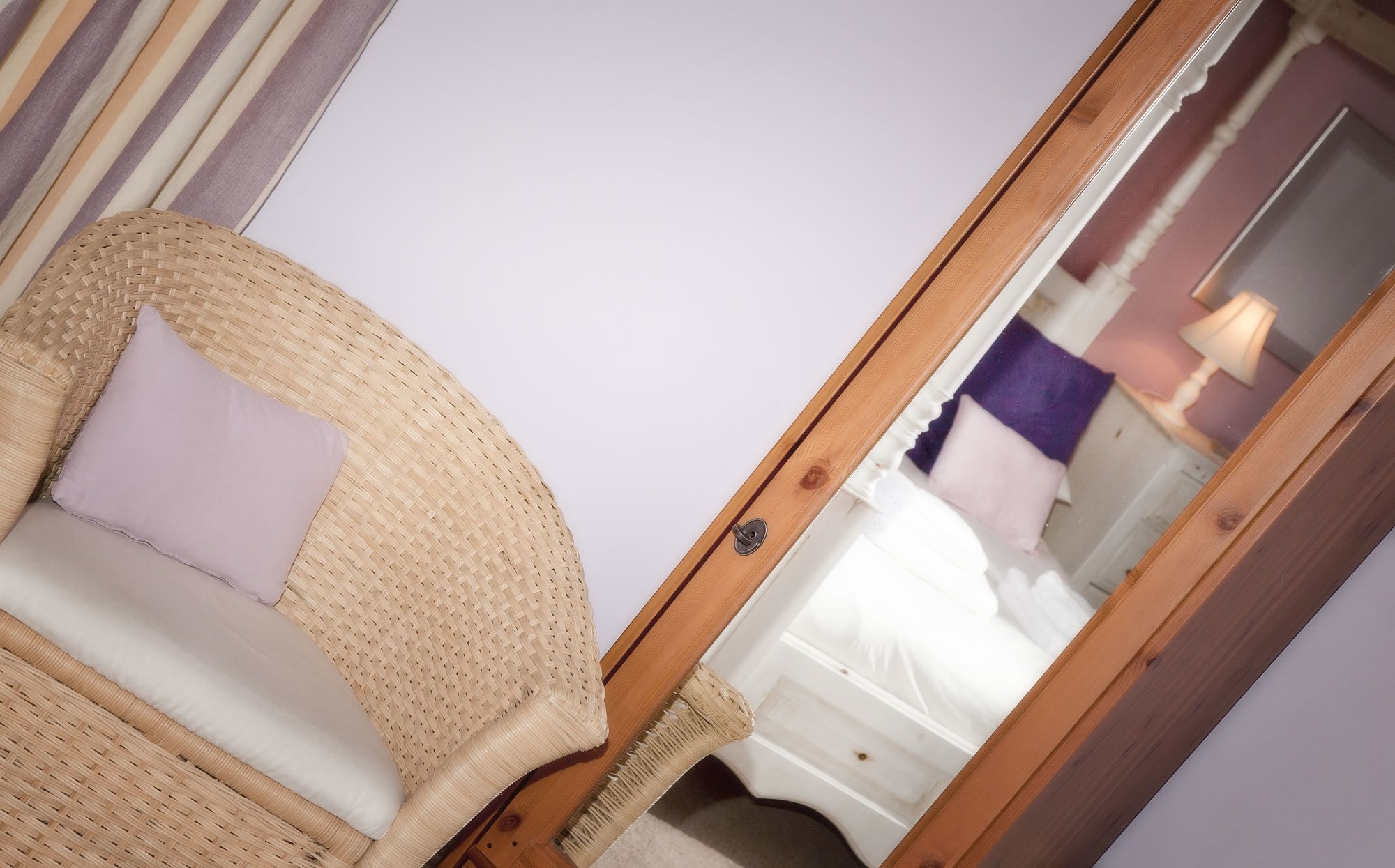 A wicker chair with a cushion in a room with a mirror reflecting a bed and a lamp.