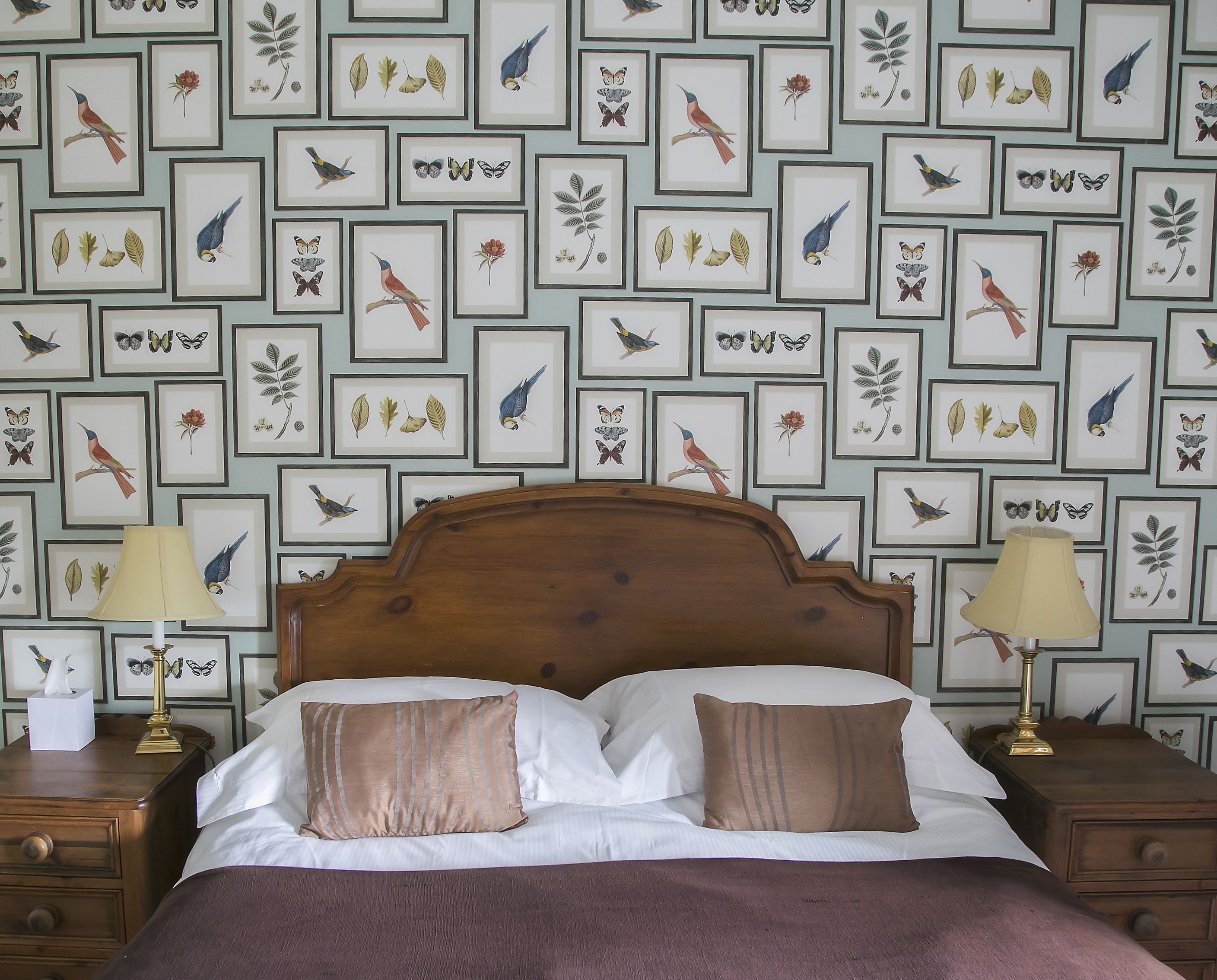 A neatly made bed with a wooden headboard, two bedside tables with lamps, and a wall decorated with framed botanical prints.