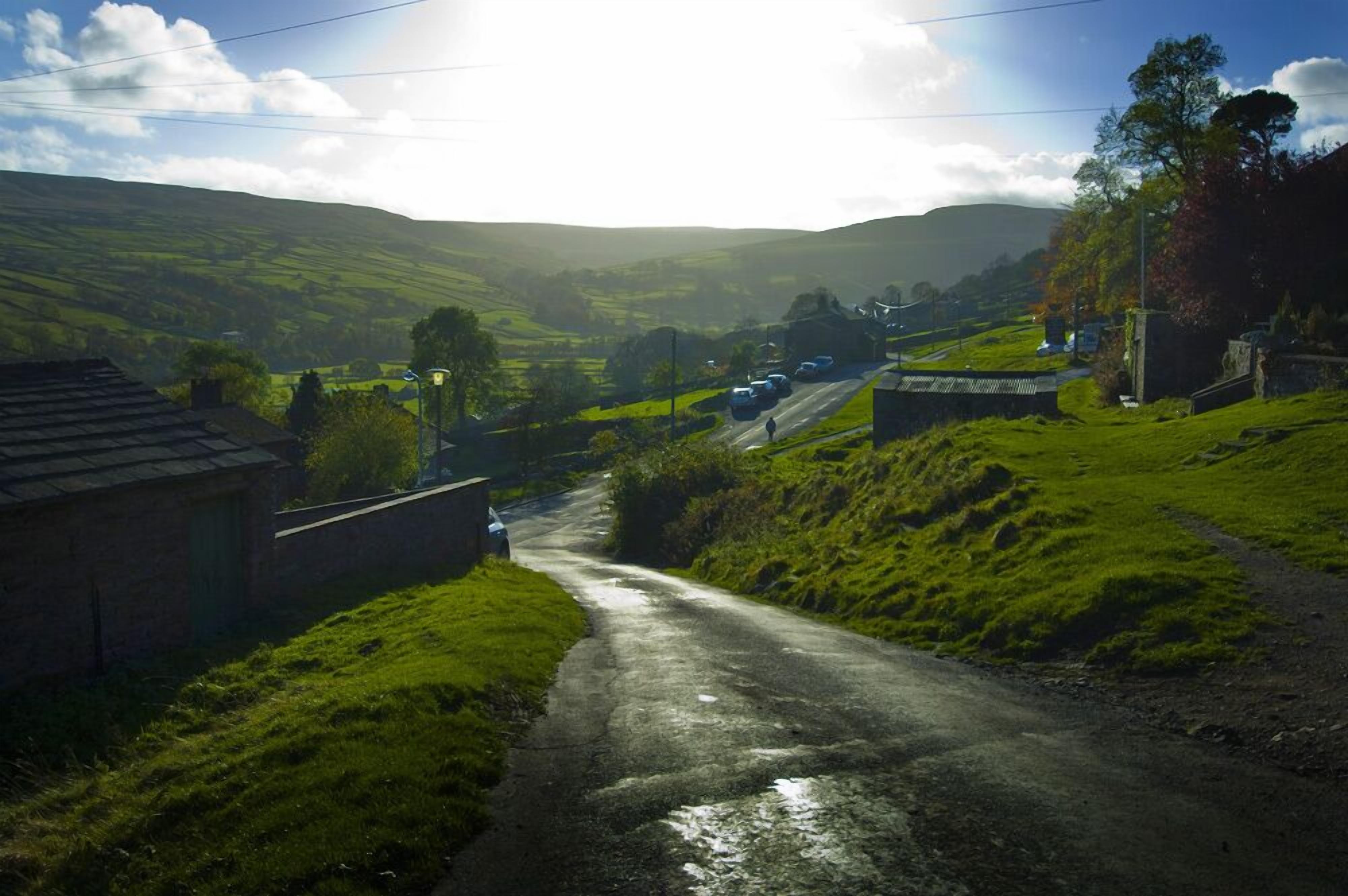 A rural road with houses on either side, surrounded by green hills.