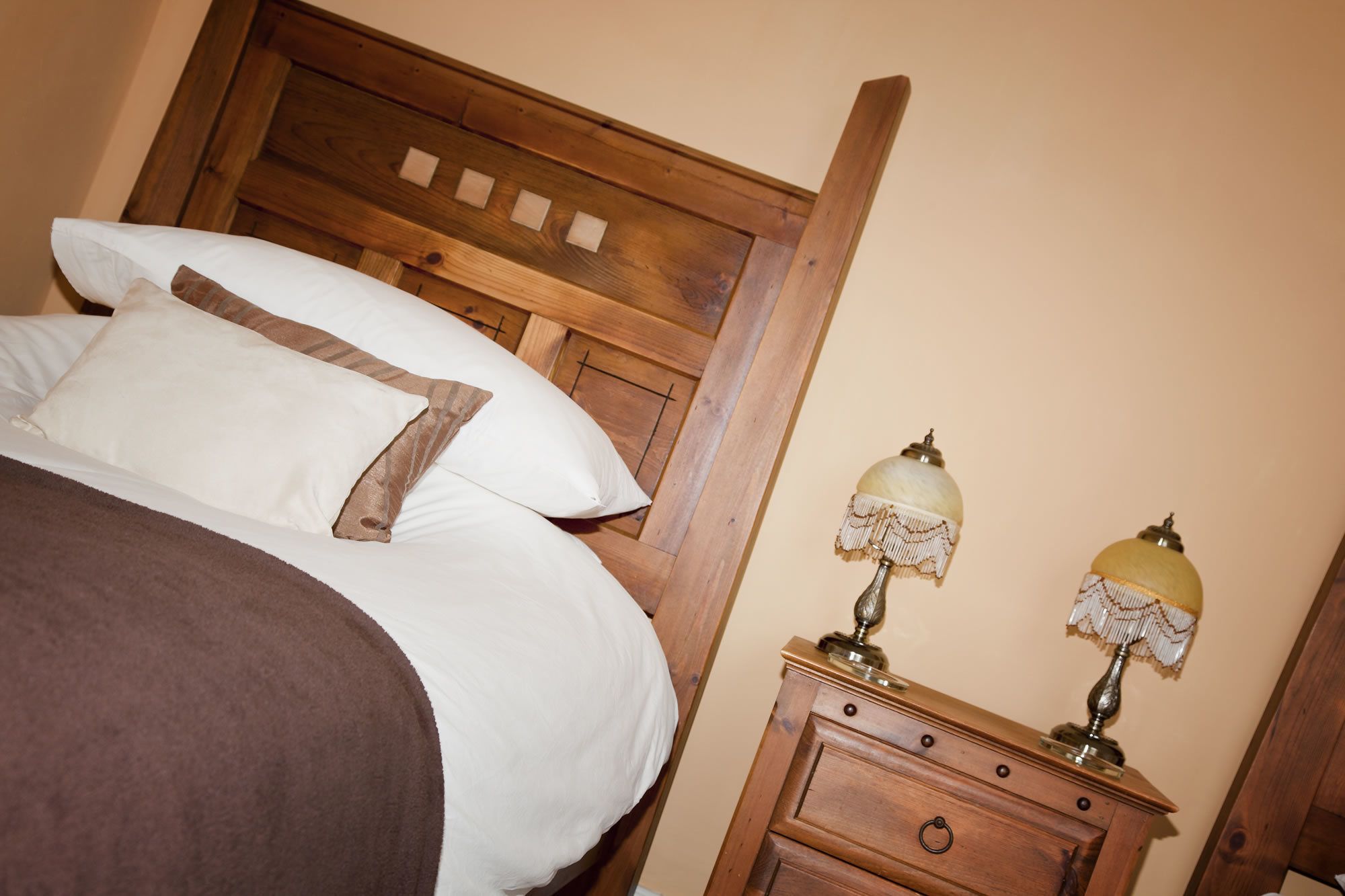 Una cama con ropa blanca y un plaid marrón, un cabecero de madera, y una mesita de noche con dos lámparas.