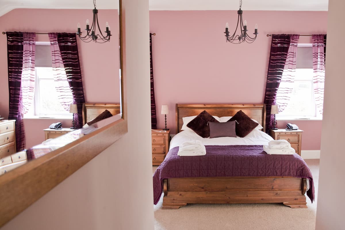 A bedroom with a wooden bed, purple bedding, a nightstand with a lamp, and a mirror reflecting the room.