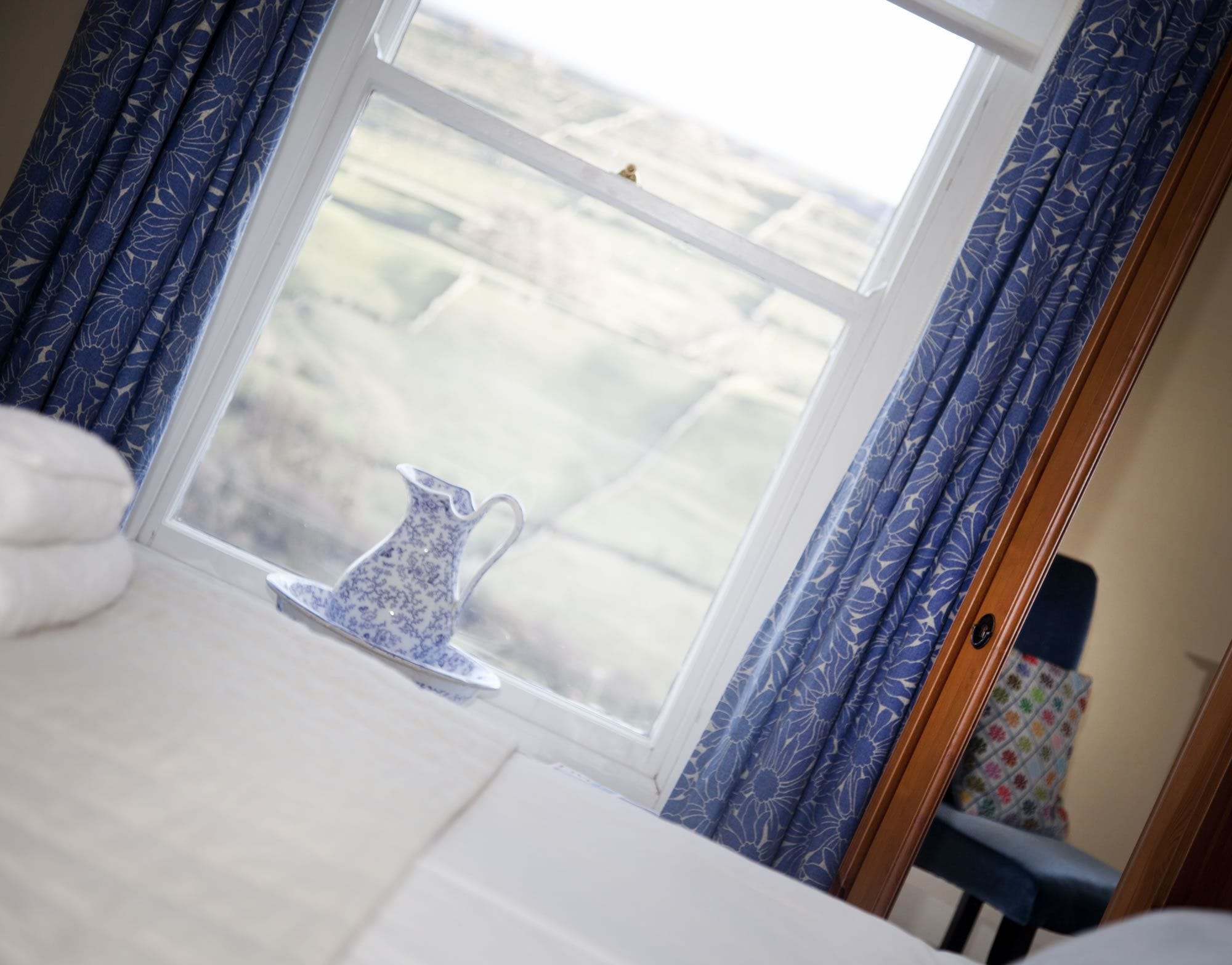 A room with a bed, a window with blue curtains, and a white pitcher on the windowsill.