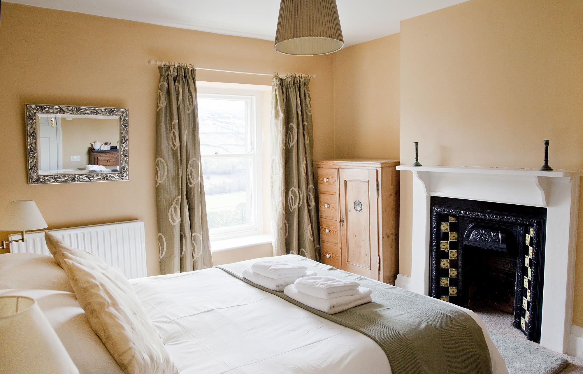 Een slaapkamer met een bed, een dressoir, een spiegel, een raam met gordijnen en een open haard.