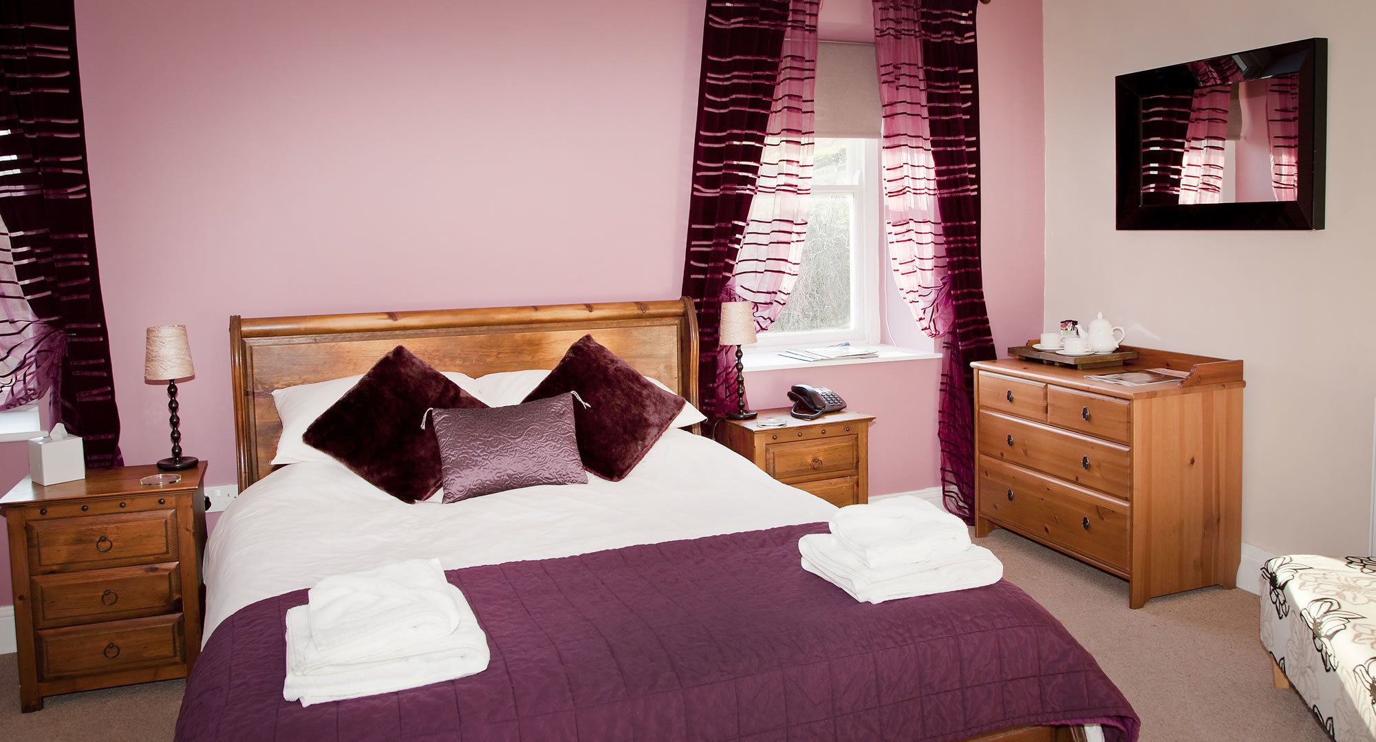 A neatly made bed with a purple bedspread and pillows, a wooden nightstand with a lamp, a wooden dresser, and a mirror on the wall.