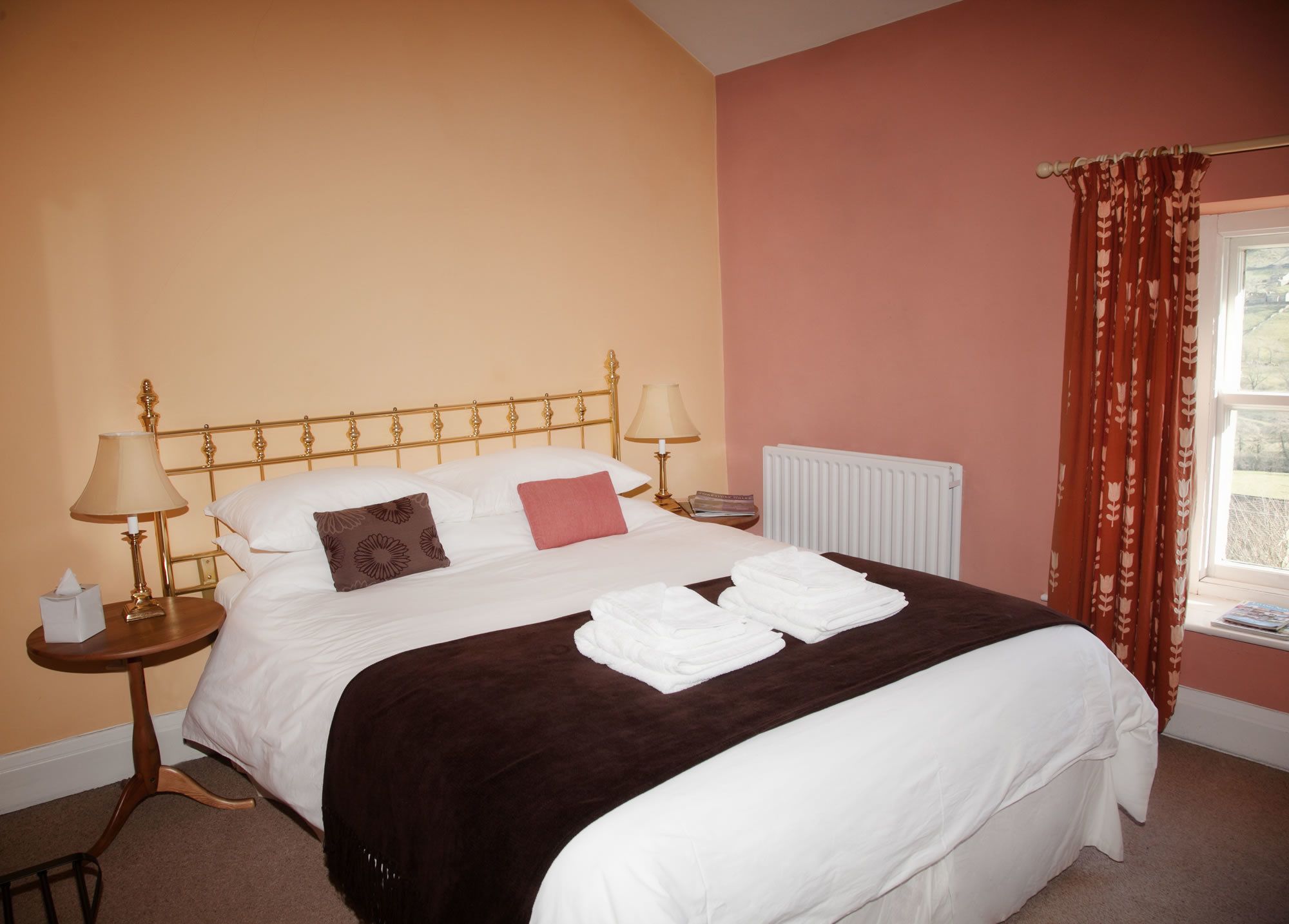 A neatly made bed with white linens, a brown throw blanket, and two pillows. There are two bedside lamps, a small table with a tissue box, and a radiator. The room has peach walls and a window with red curtains.