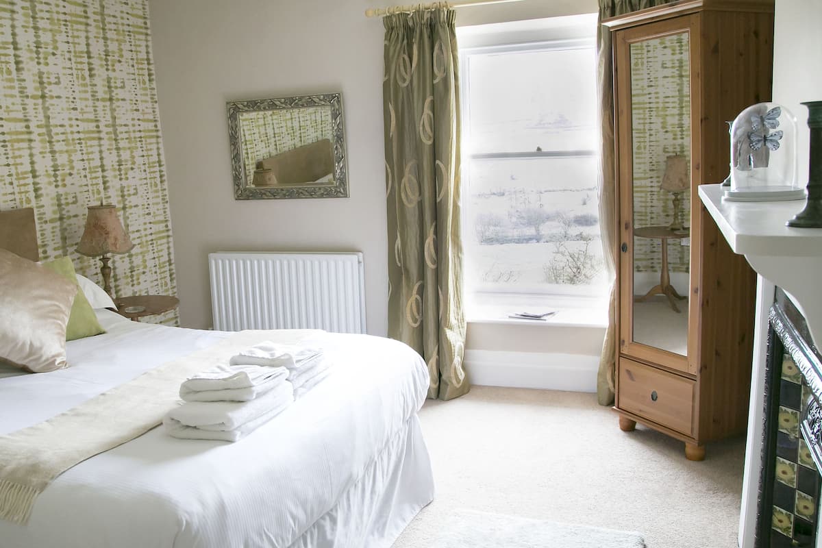 A neatly made bed with pillows, a radiator, a window with curtains, a mirror, and a cabinet.