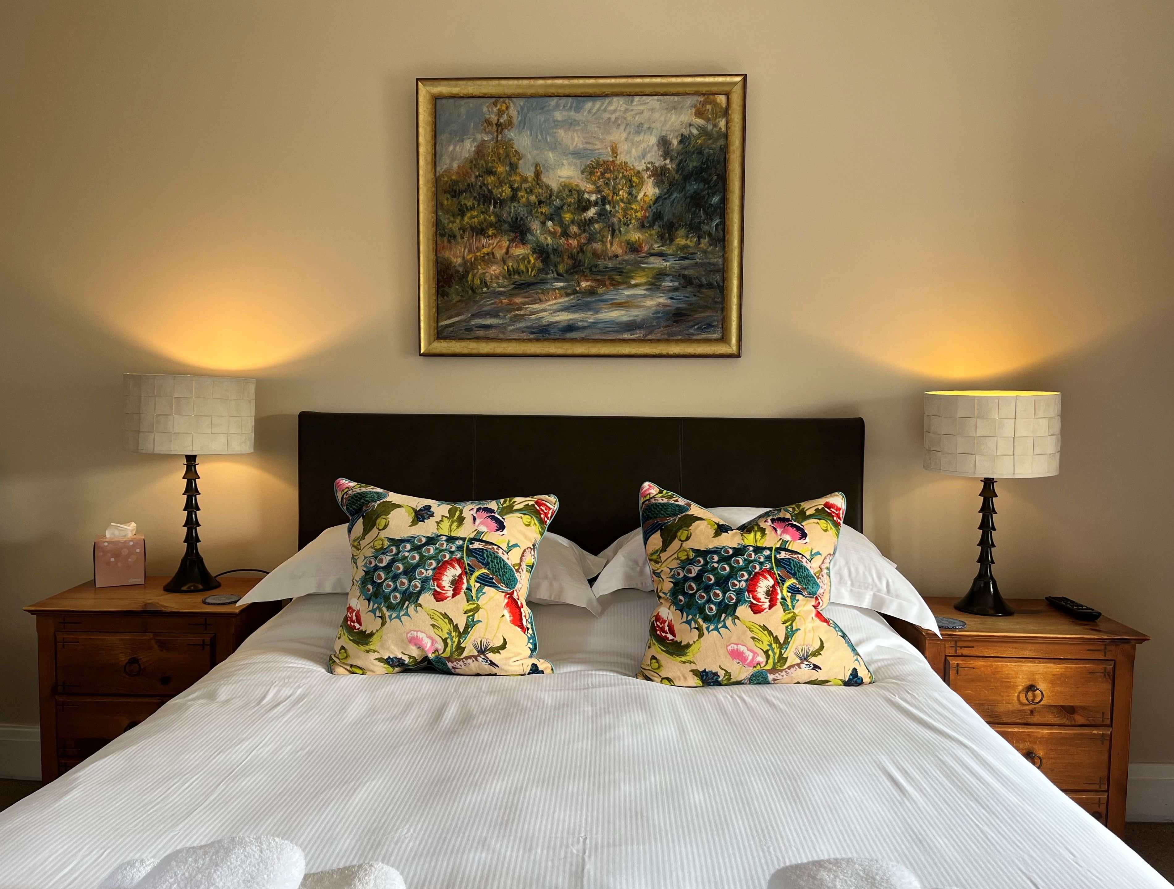 A neatly made bed with two decorative pillows, a painting above, and two bedside lamps on wooden nightstands.