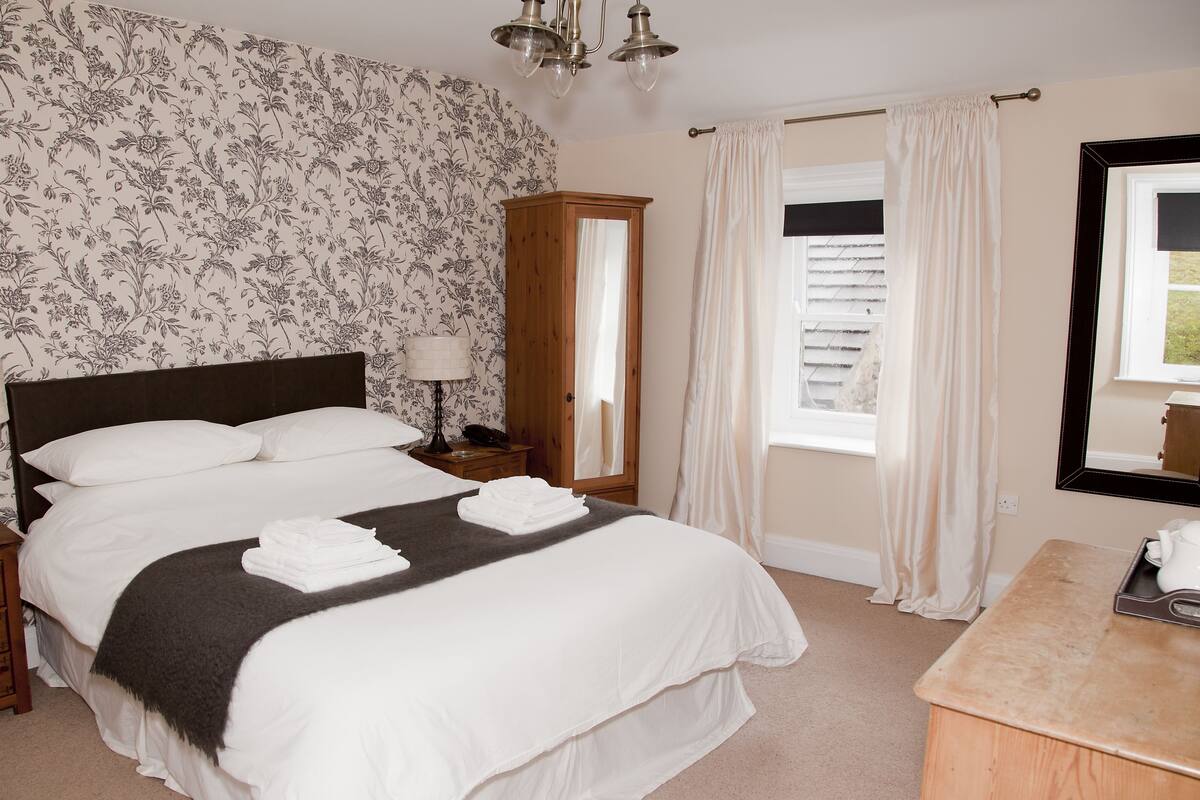 A neatly made bed with white linens, a wooden nightstand with a lamp, a mirror, and a window with curtains.