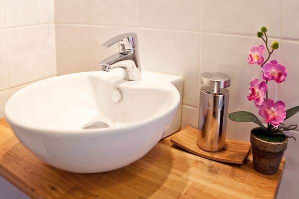 Un lavabo de cerámica blanca con un grifo moderno, una botella de acero inoxidable y una pequeña orquídea en maceta sobre una encimera de madera.