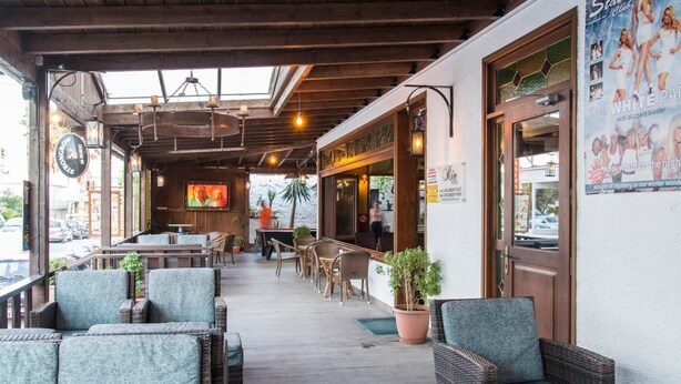 An outdoor seating area with wicker chairs and tables, a wooden deck, and a view of the street. - Rio Hotel (Kos)