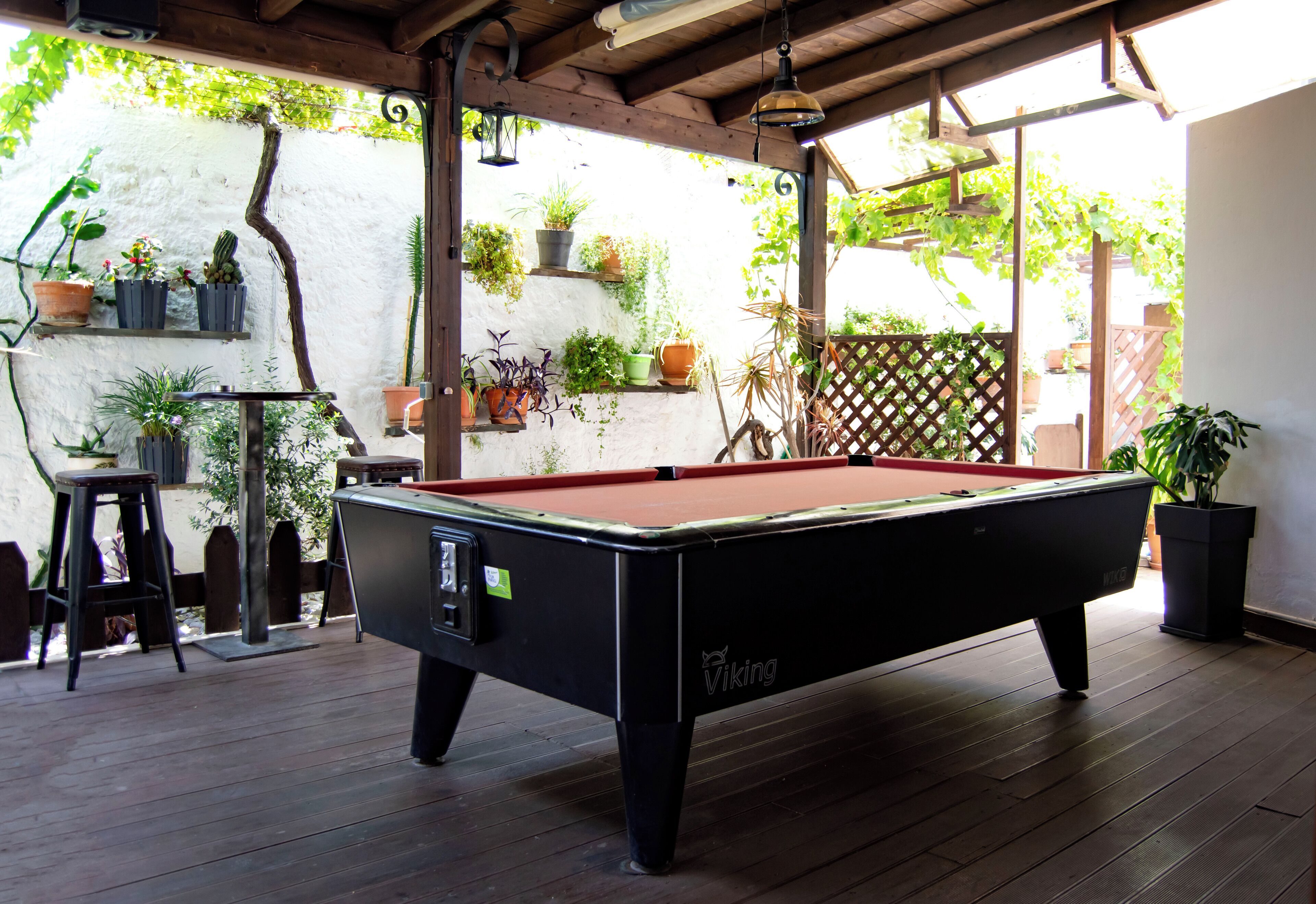 Une table de billard se trouve dans un espace extérieur abrité, doté d’un plancher en bois et agrémenté de plantes en pot.