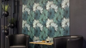 A modern room with a patterned wall, two black leather chairs, a wooden table with a croissant and a cup of coffee, and potted plants on a black metal stand.